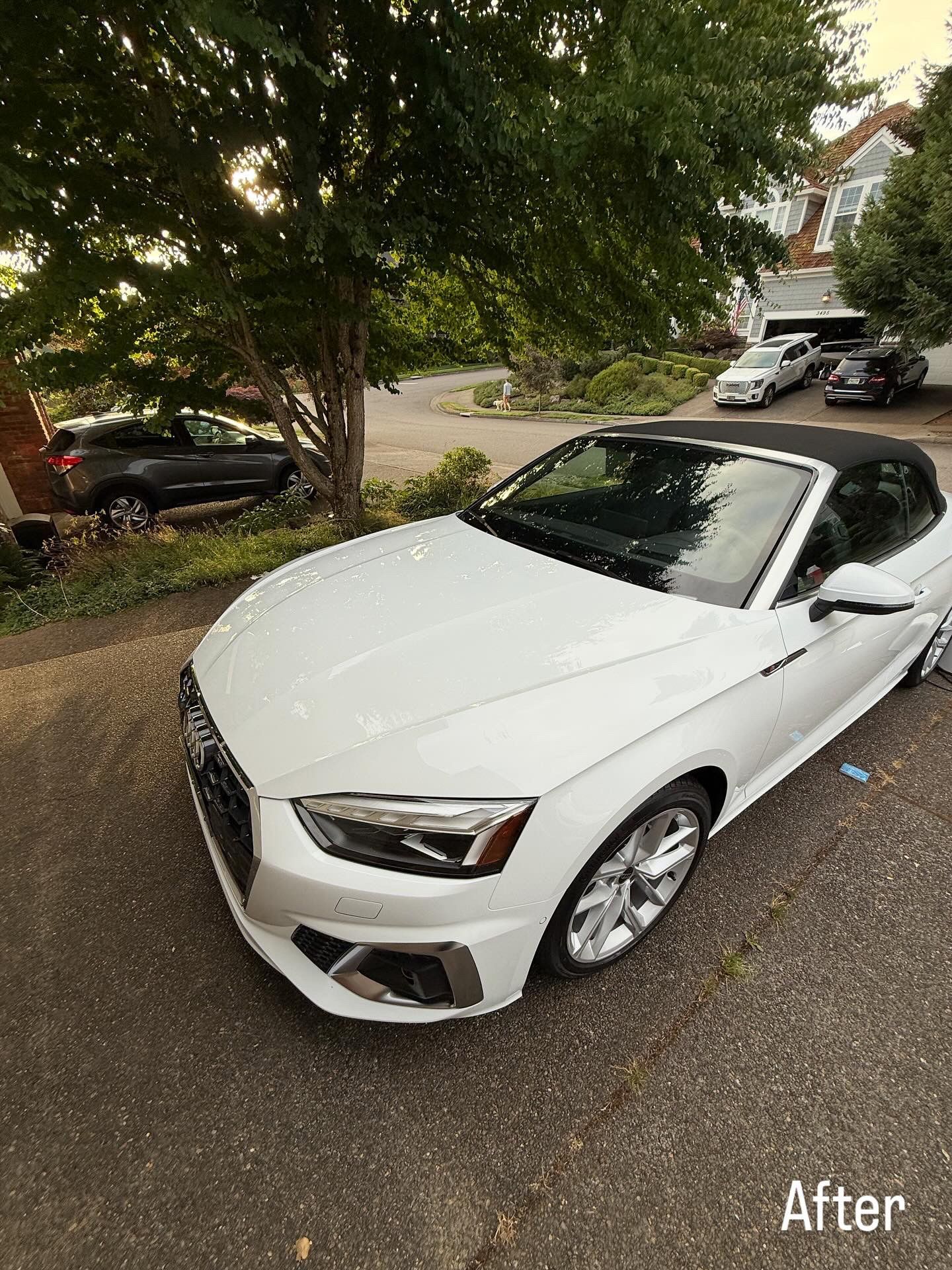 White convertible car parked on a driveway under trees, with a residential street in the background.