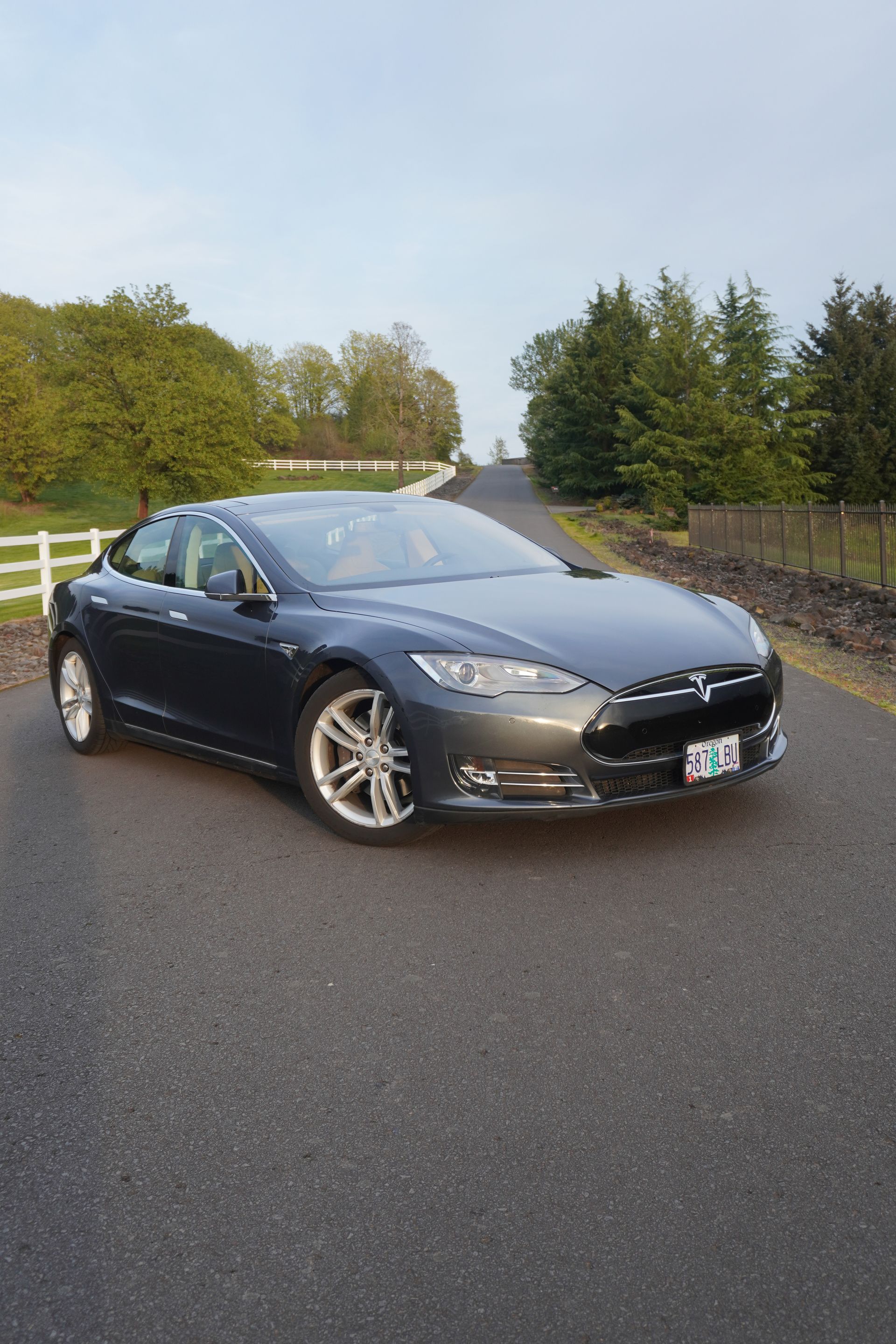 Dark blue sedan parked on a rural road beside trees and a white fence