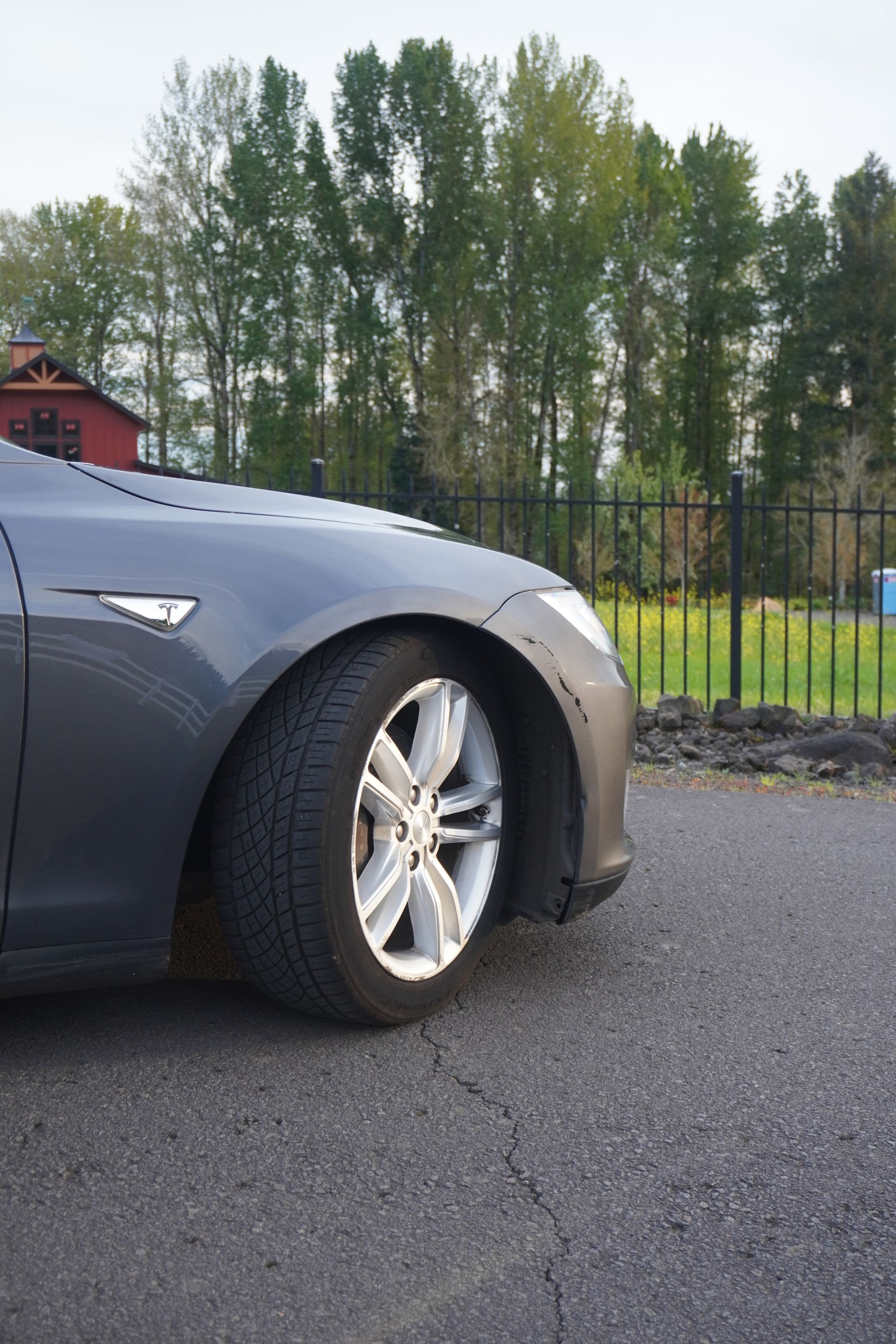 Front of a gray car parked on asphalt near a fence with trees in the background