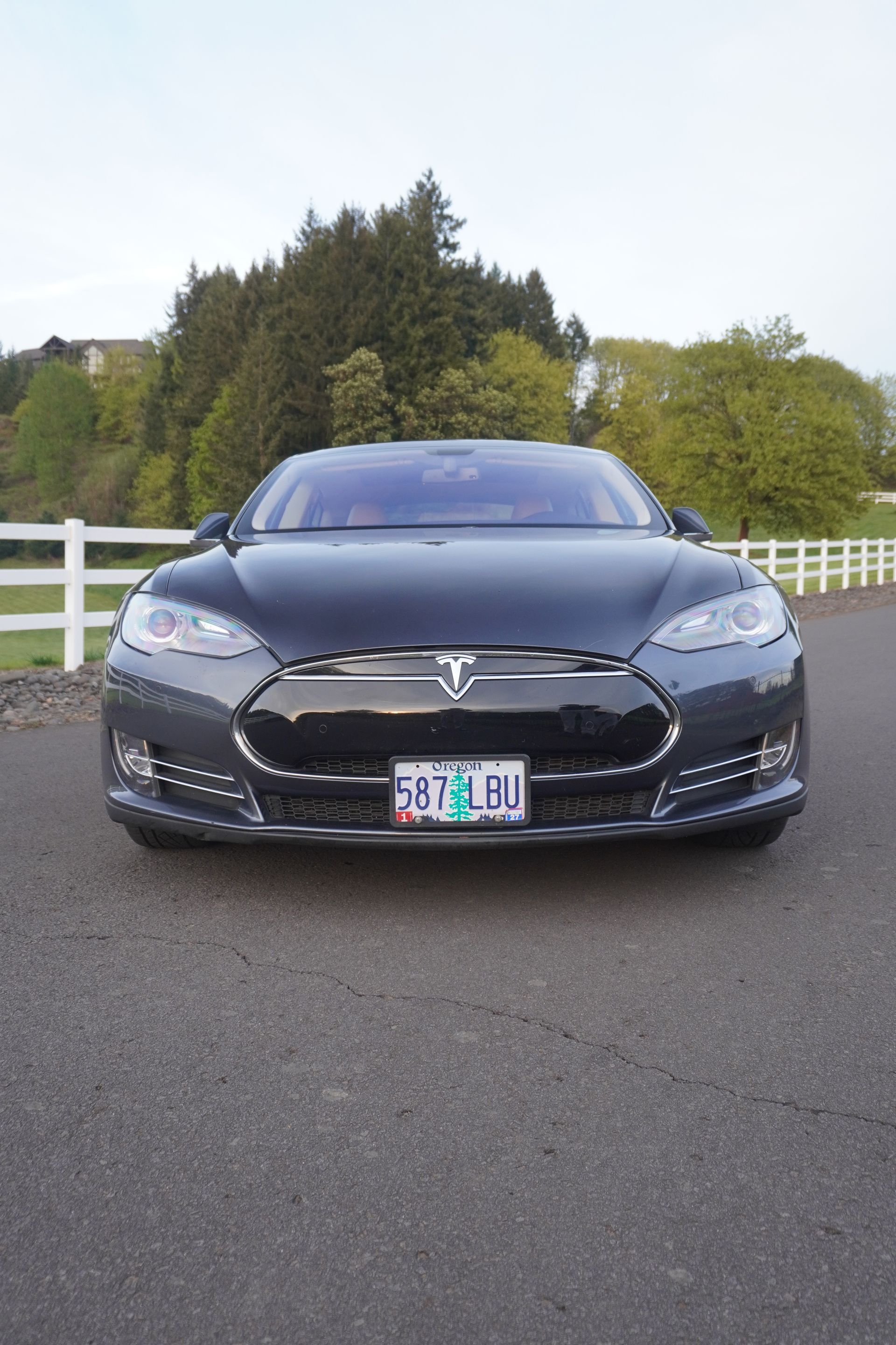 Front view of a black Tesla parked on a road with trees and a white fence in the background