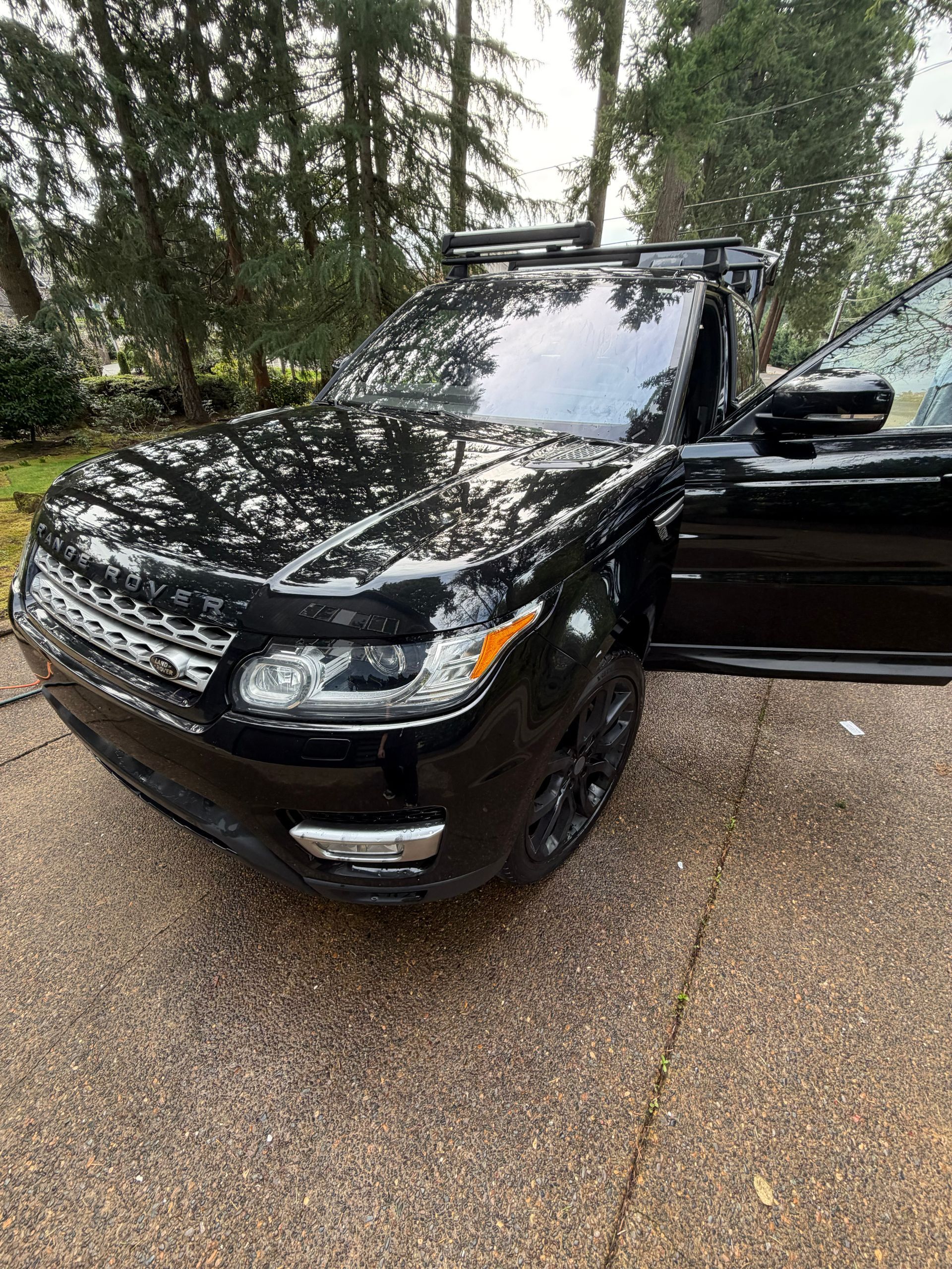 Black SUV parked on a wet driveway with the driver’s door open, trees in the background