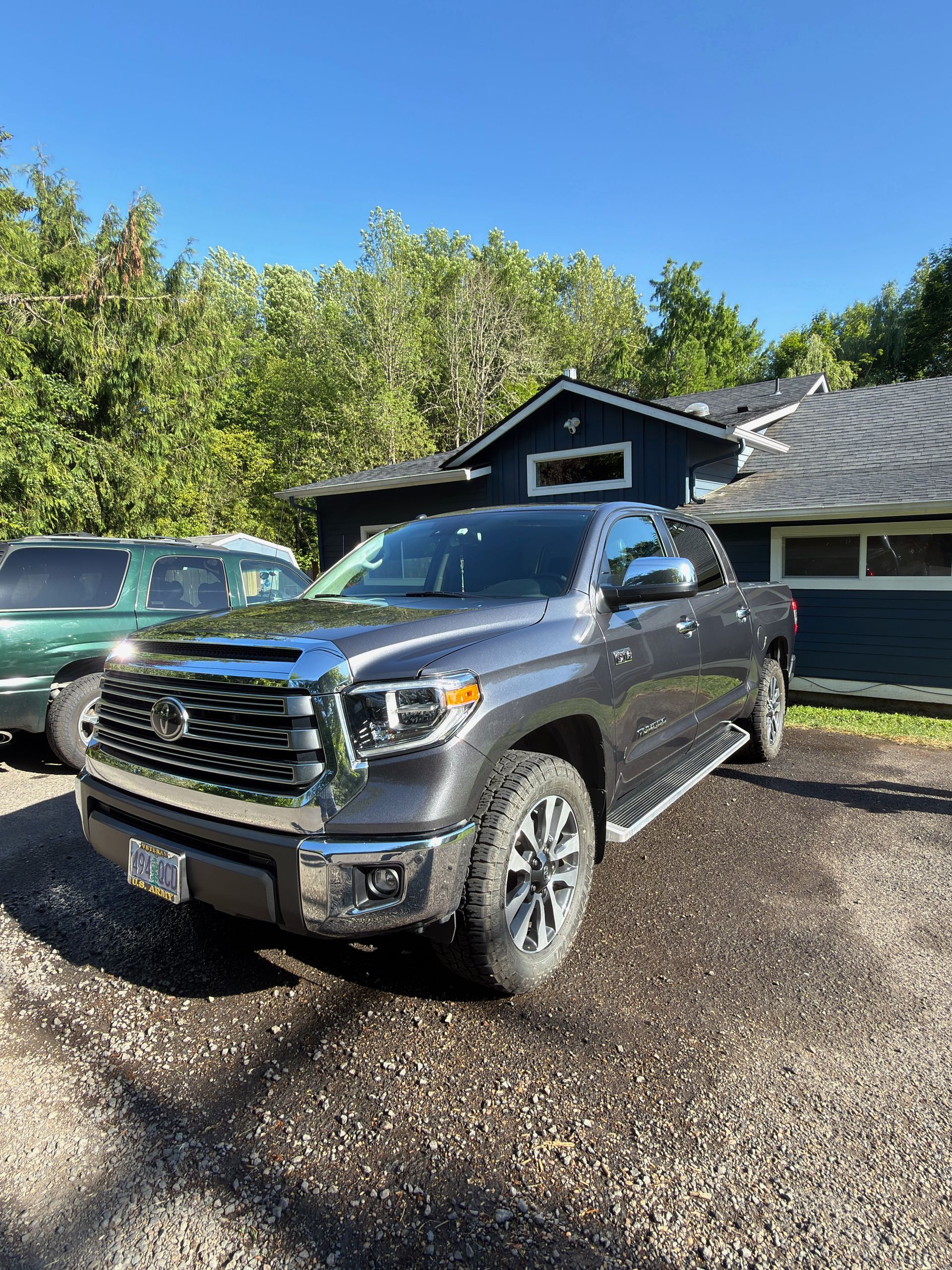 Gray pickup truck parked in a driveway beside a green SUV and a house on a sunny day