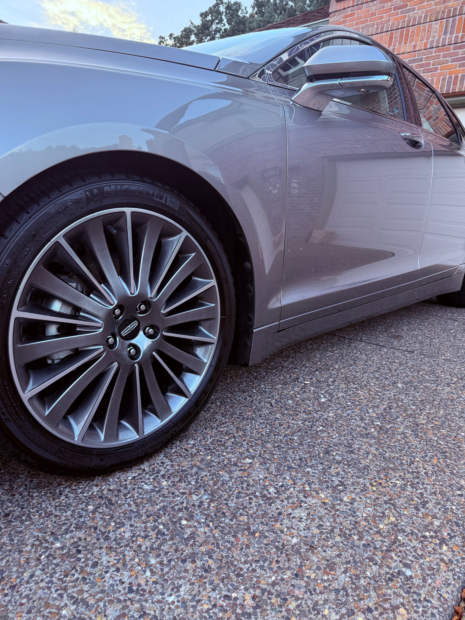 Close-up of a silver sports car’s front wheel and side door on a gravel surface.