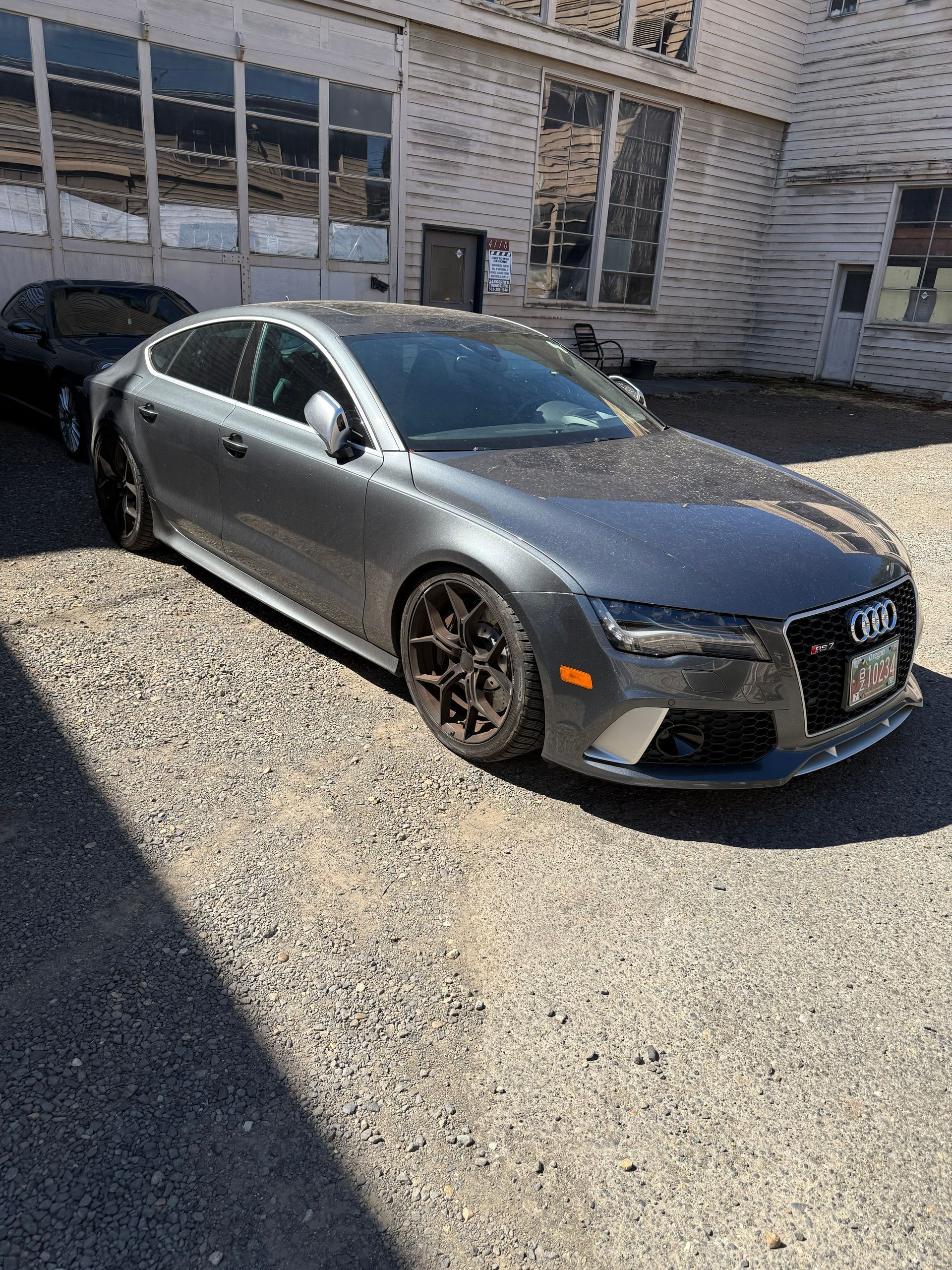 Gray sedan parked on gravel outside an industrial building, with a black car behind it.