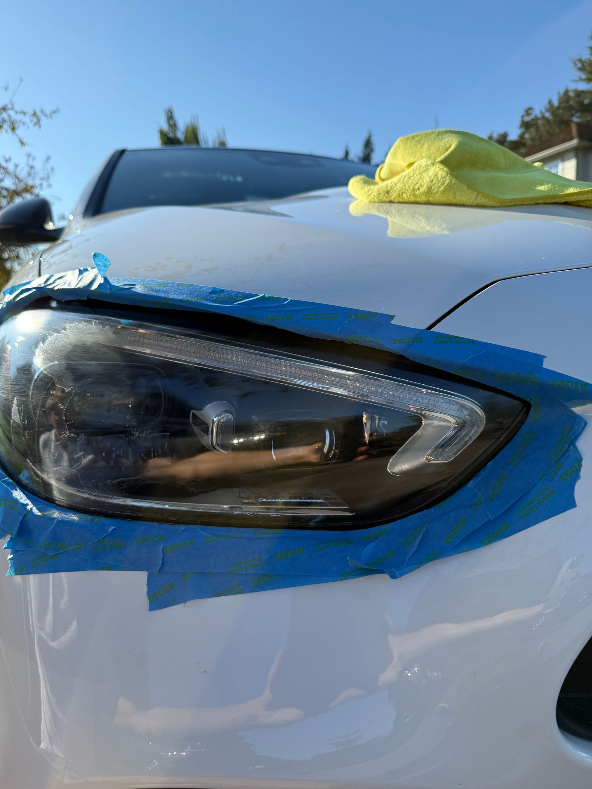 Close-up of a car’s front headlight and hood with blue tape and a yellow cloth on top