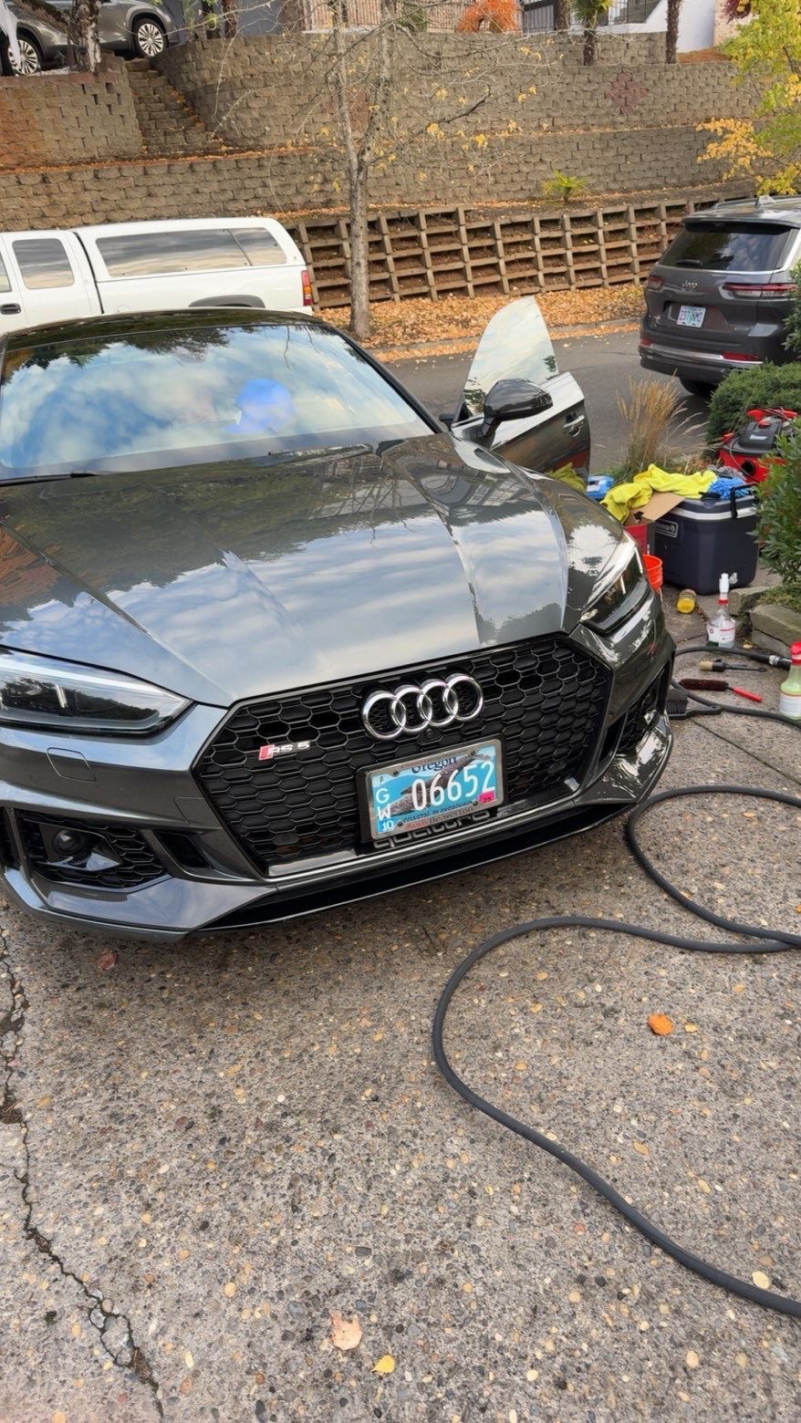Black Audi sedan being washed outdoors with hose and cleaning supplies in a driveway