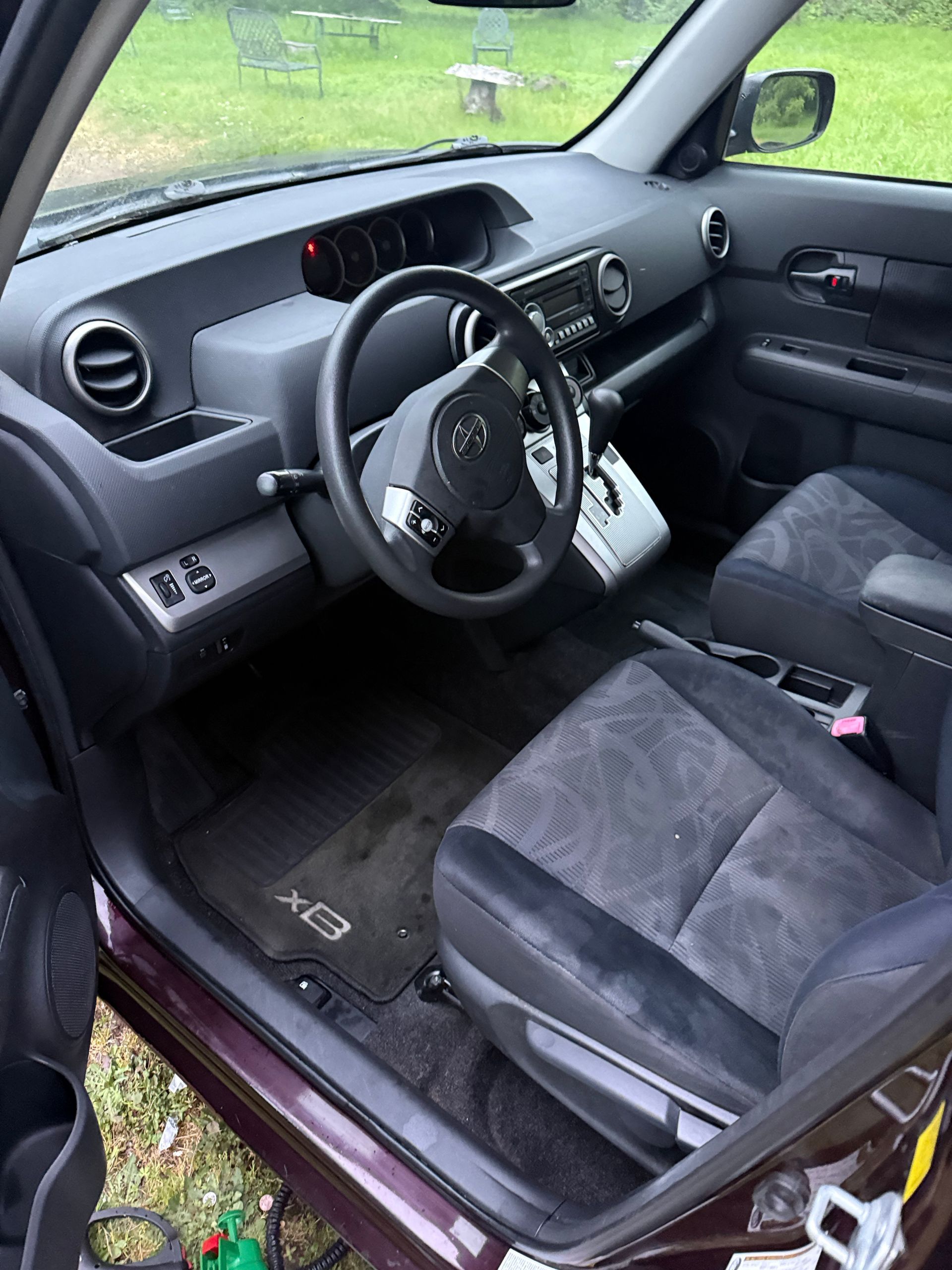 Driver’s side interior of a car with black seats, steering wheel, and dashboard, door open to a grassy outdoors