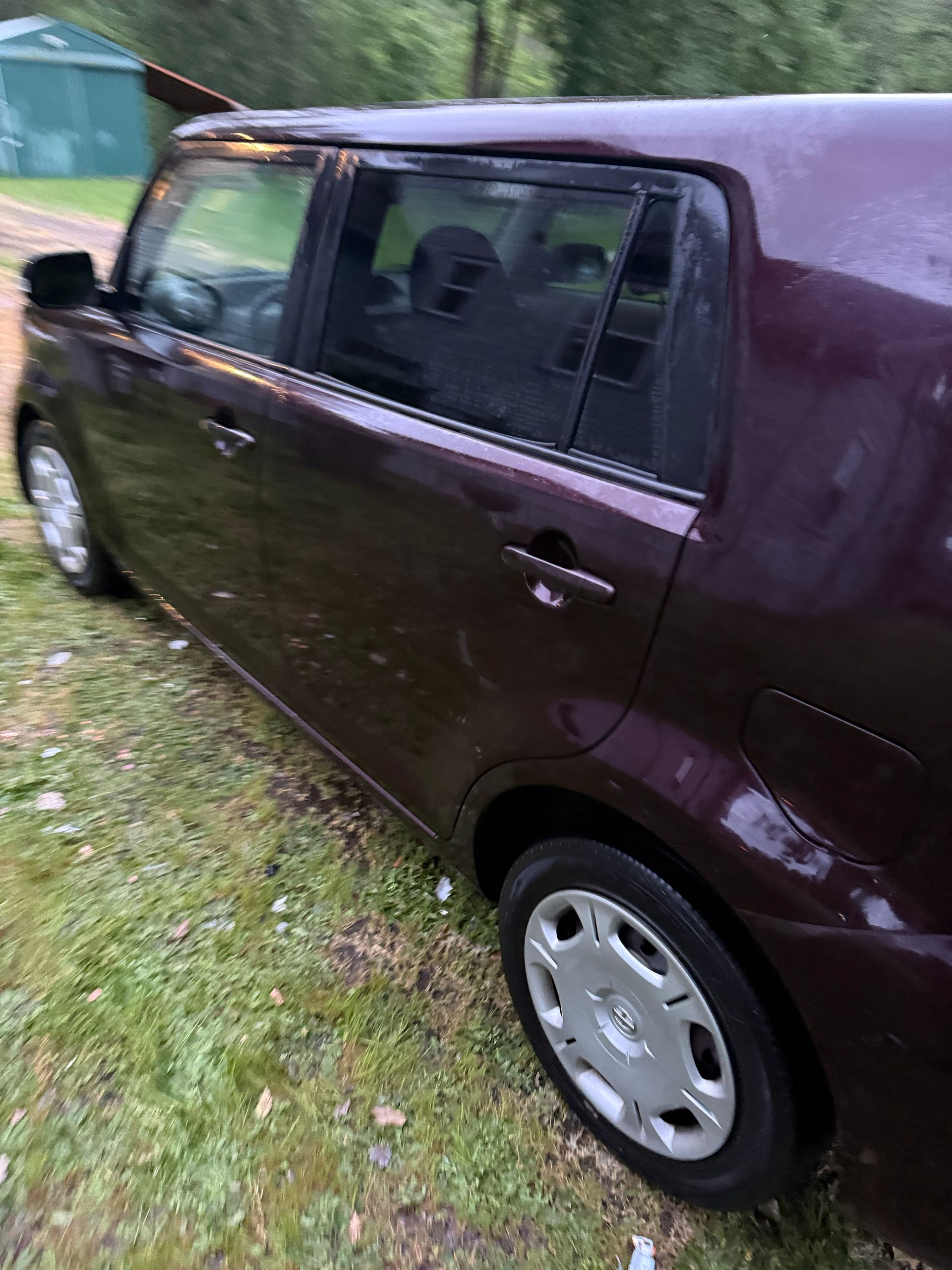 Dark maroon hatchback parked on grass beside a road, viewed from the side.