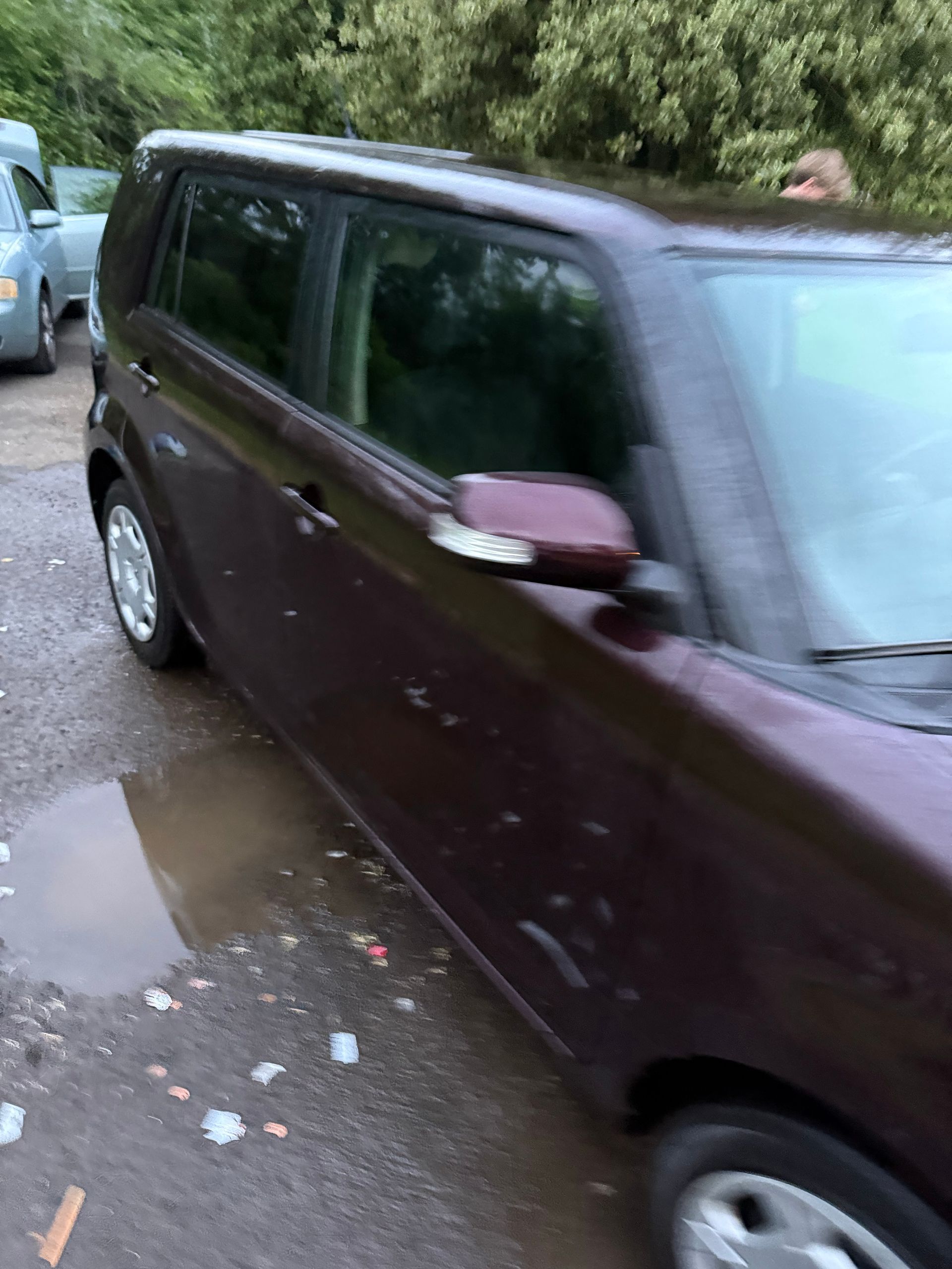 Maroon minivan driving on a wet road through a wooded area, with another car behind it.
