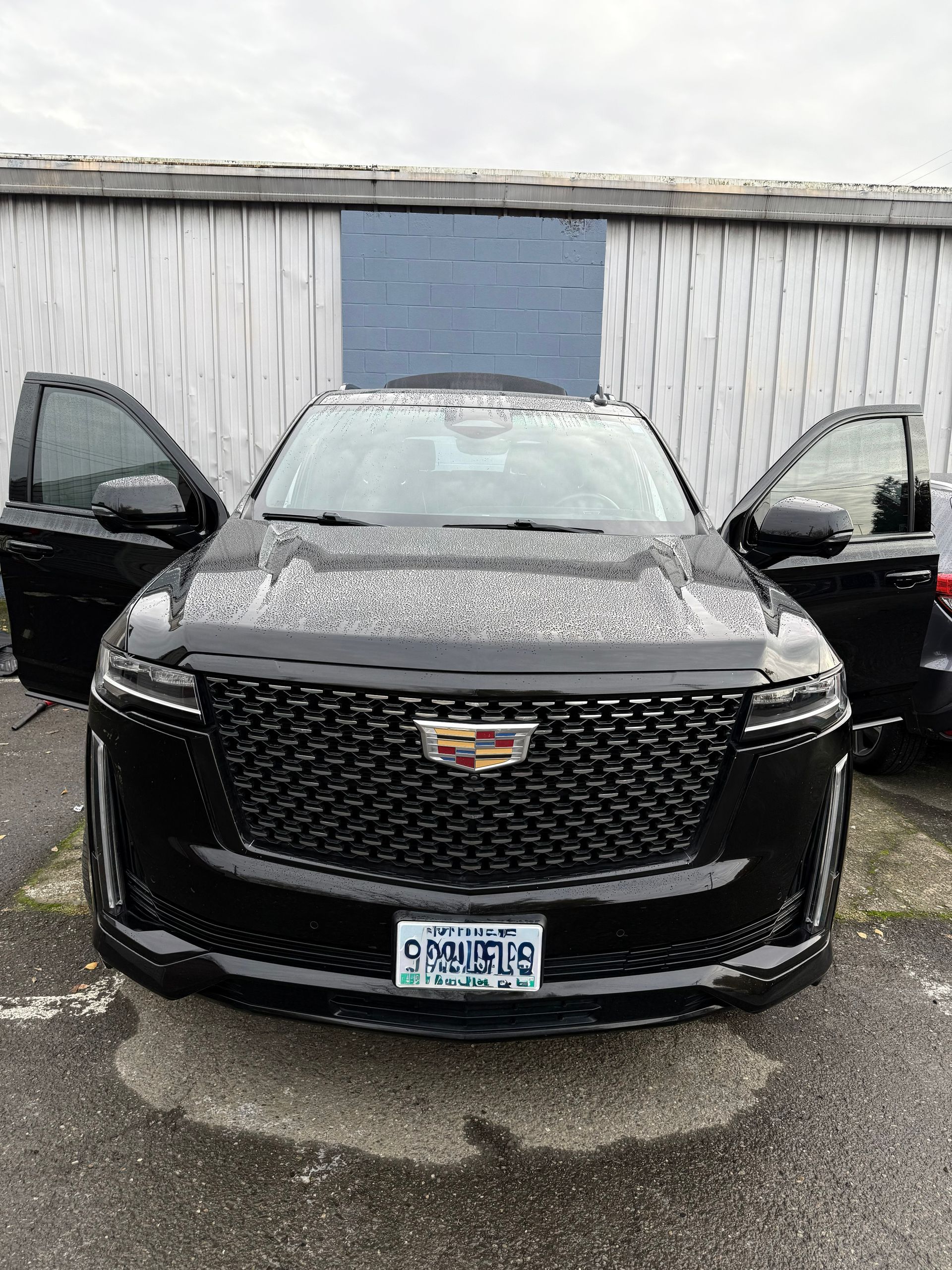 Black Cadillac SUV parked with both front doors open in front of a garage on a wet lot.