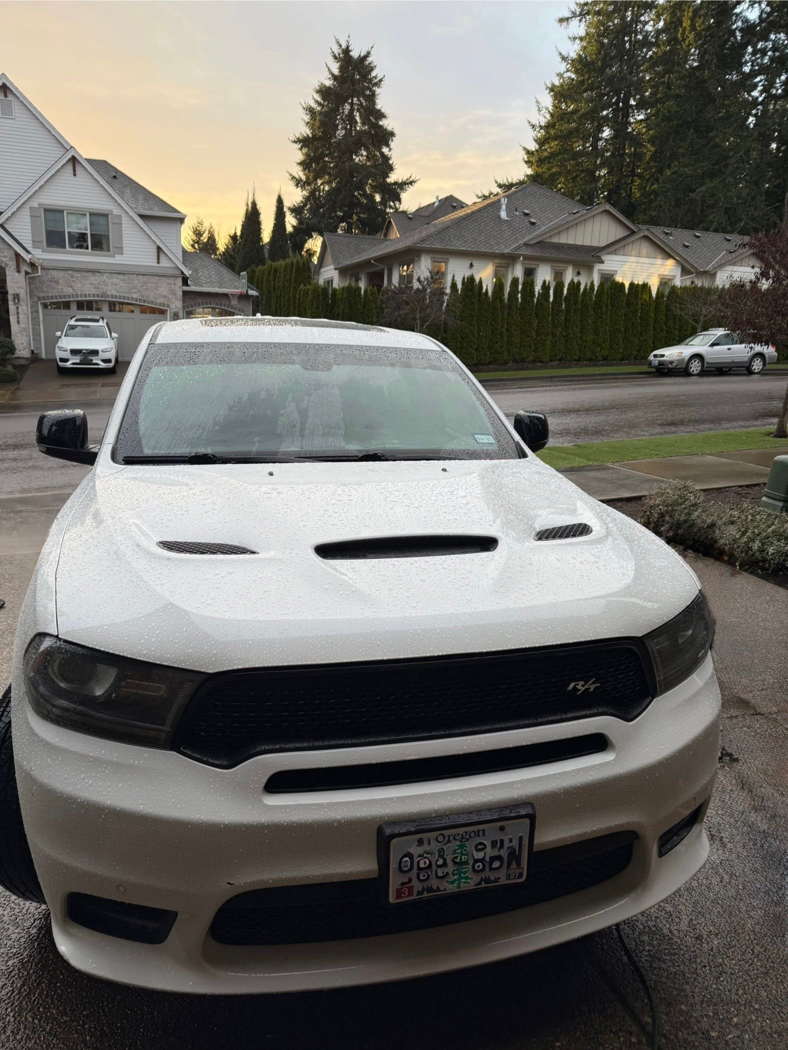 Snow-covered white SUV parked in a suburban driveway at dusk, with houses and trees in the background.