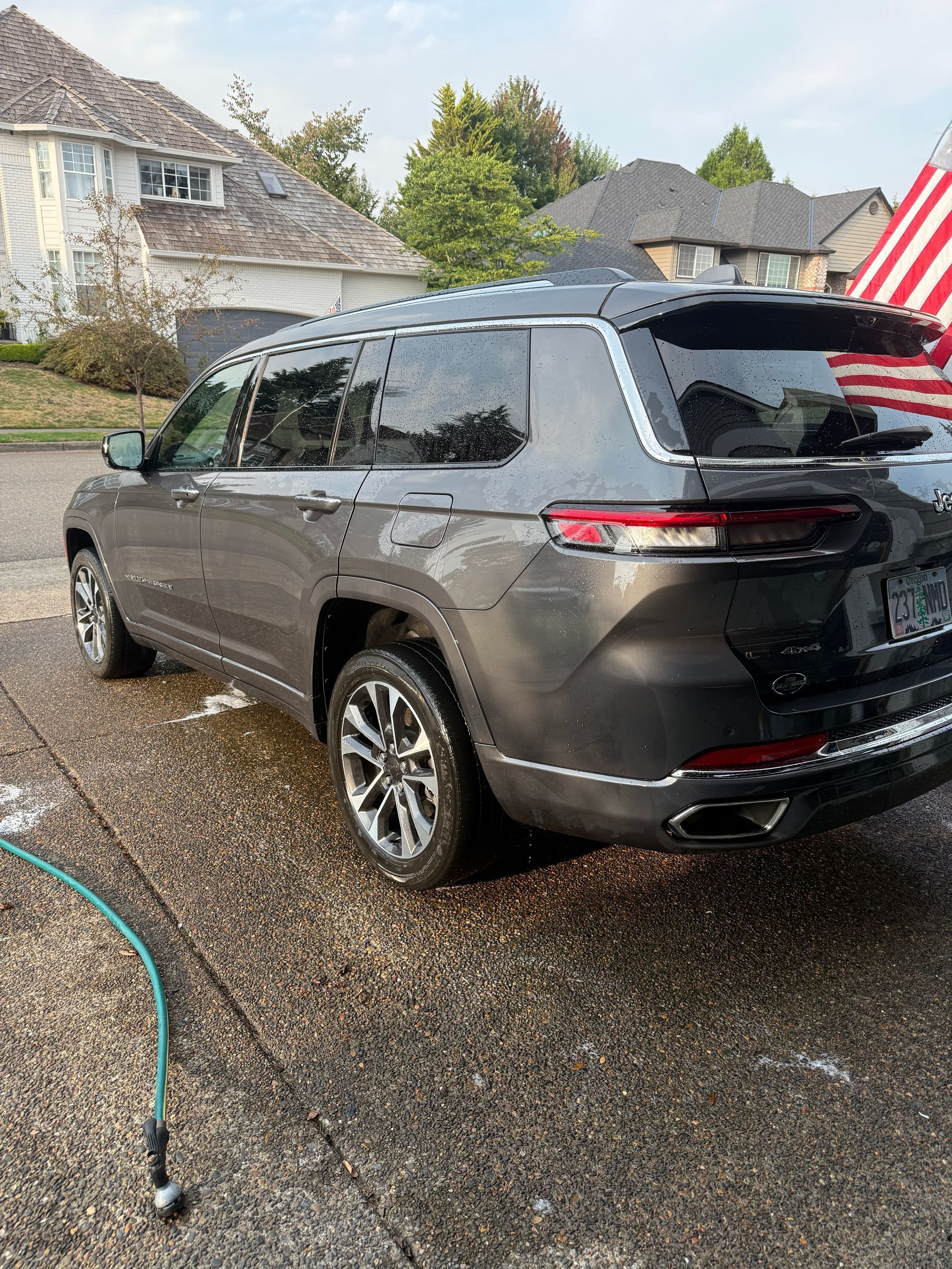 Dark gray SUV parked on a wet driveway in front of suburban houses, with an American flag visible.