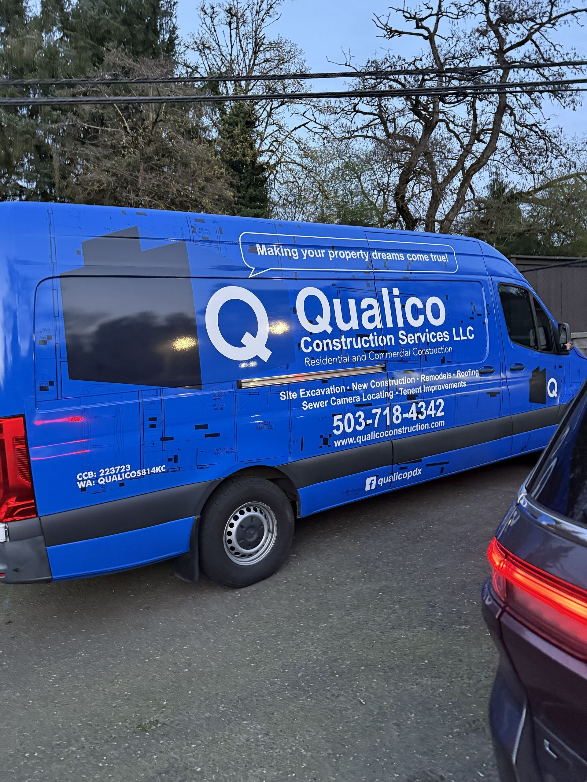 Blue Qualico Commercial Services van parked on a street beside a car and trees.