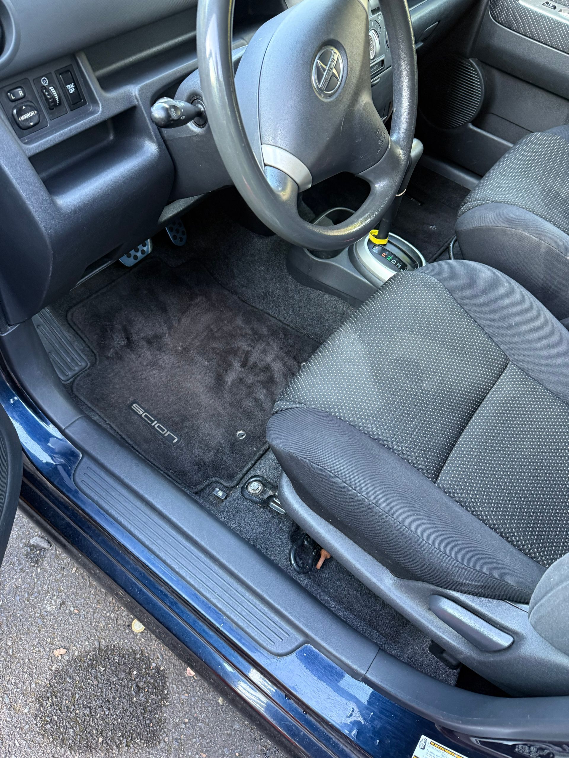 Car interior with worn black seats and dark floor mat, viewed from the open driver’s door