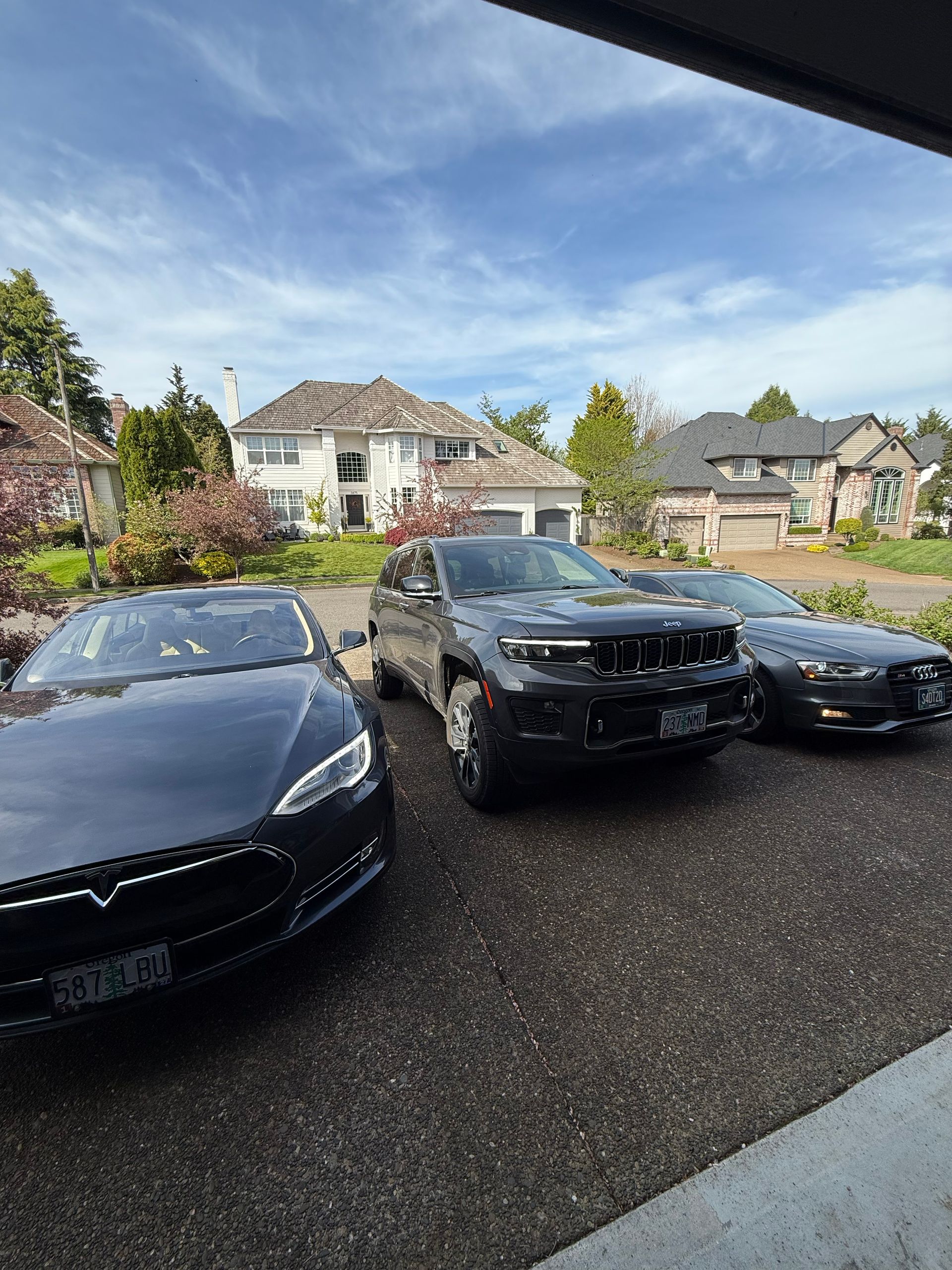Three cars parked in a driveway on a suburban street under a partly cloudy sky.