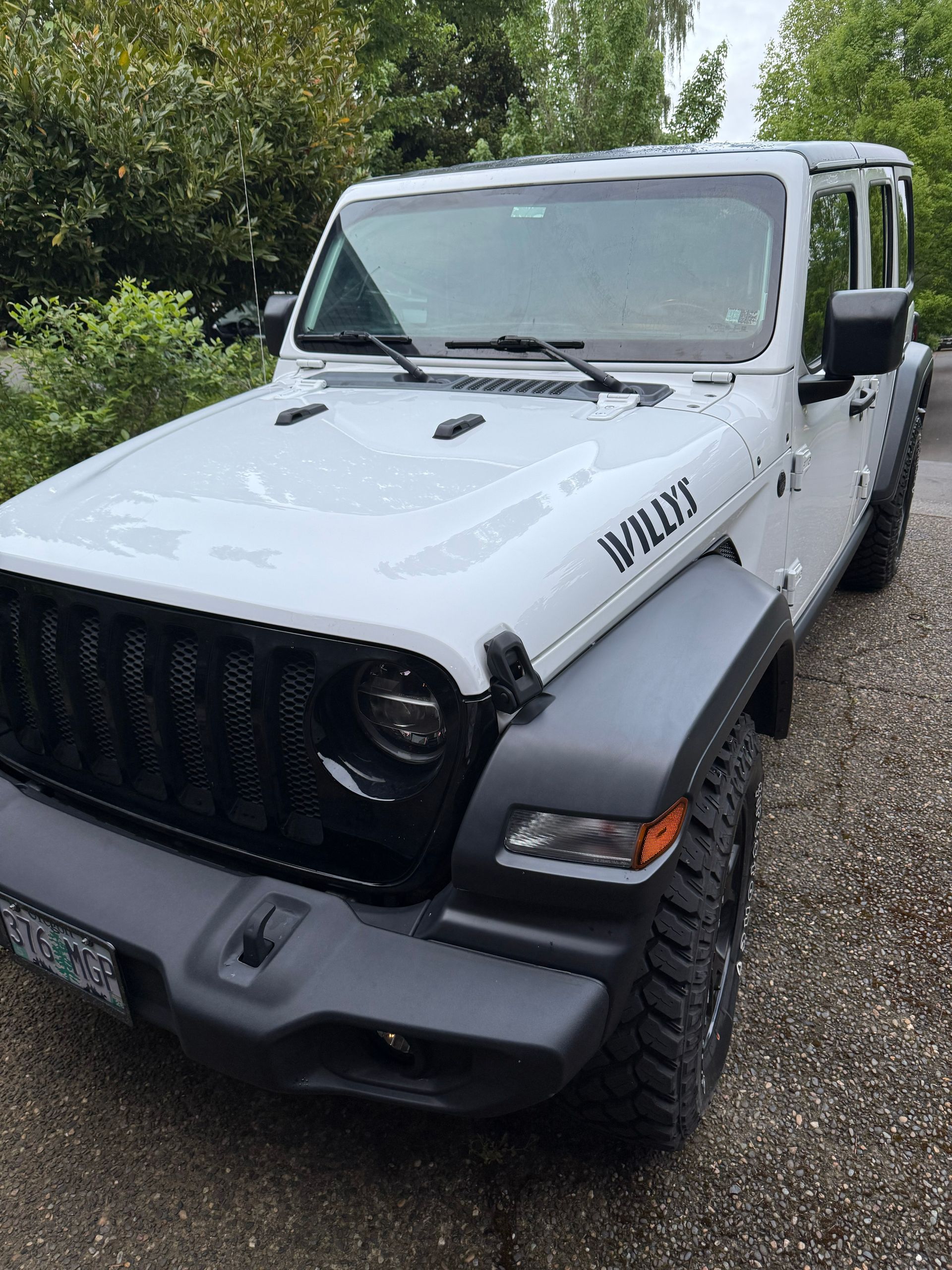 White Jeep Wrangler parked outdoors on a gravel driveway, front-left view with black grille and fenders