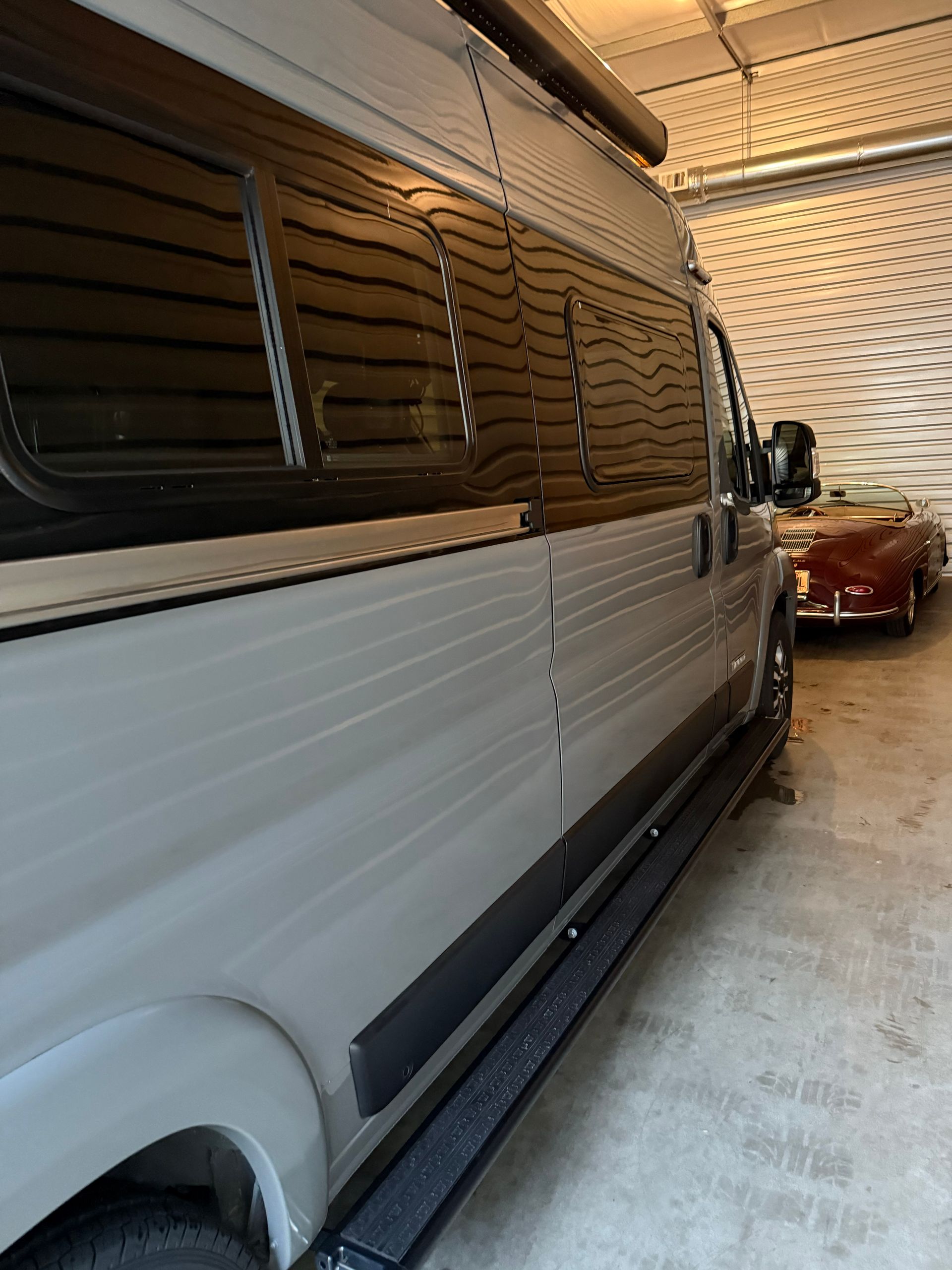 White van in a narrow indoor garage beside a red car and perforated metal wall.