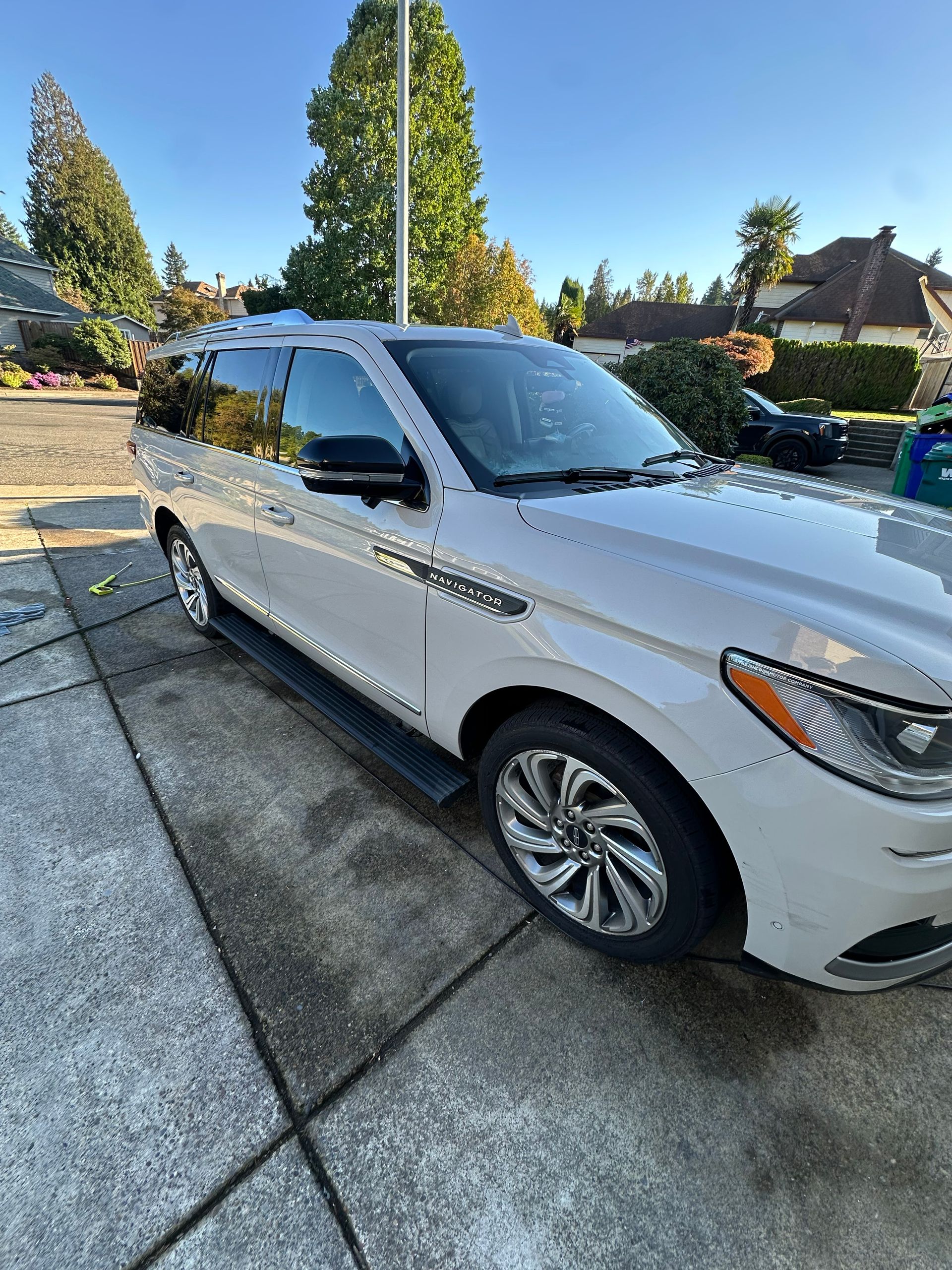 White SUV parked in a residential driveway beside a sidewalk, with trees and houses in the background.
