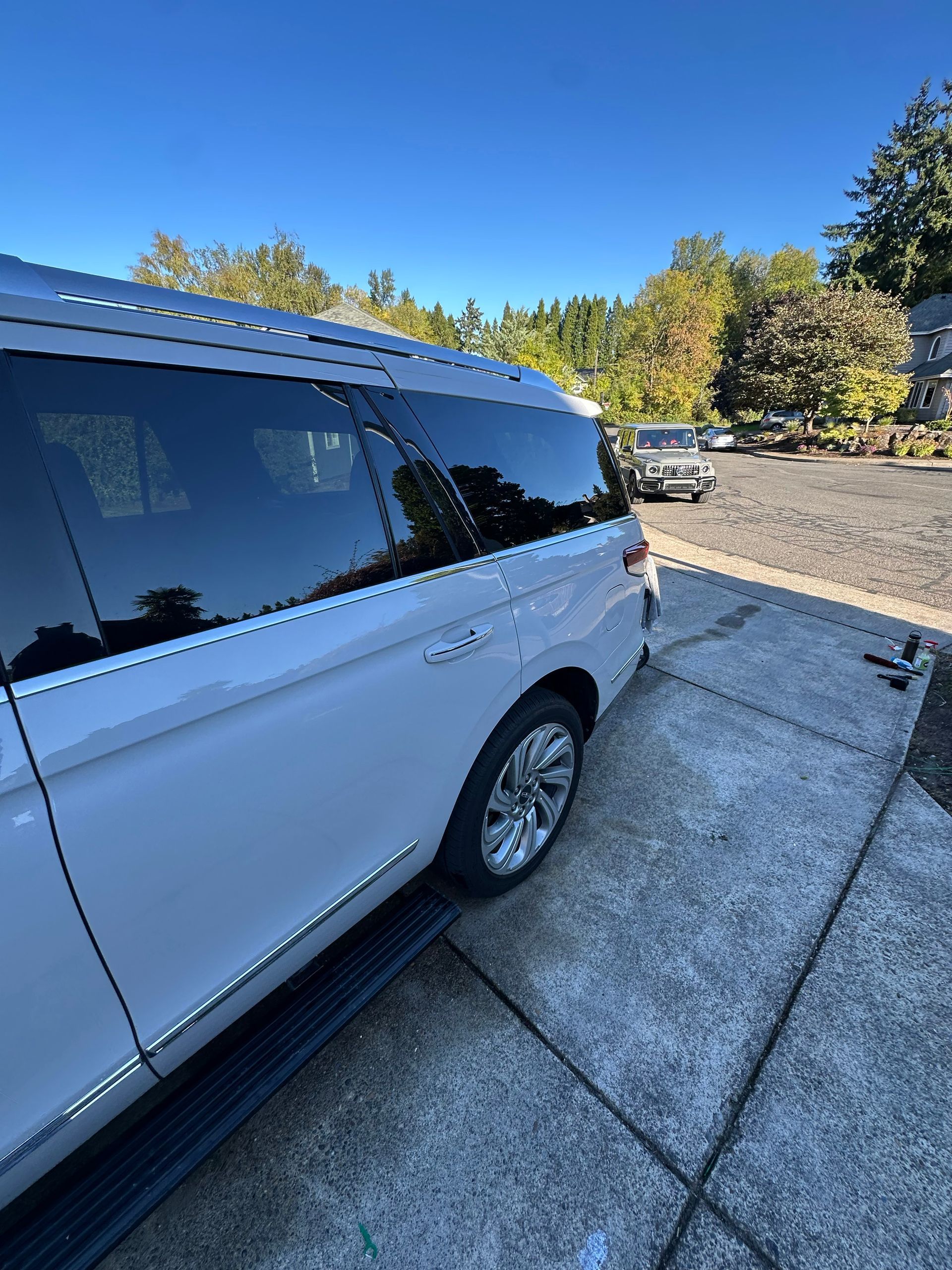 White SUV parked beside a concrete driveway on a sunny day, with trees and another car in the background