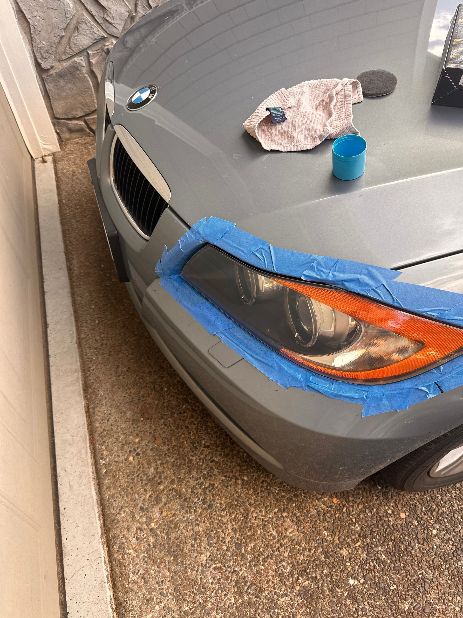 Silver car hood with cleaning supplies and blue tape around the headlight, parked outdoors.