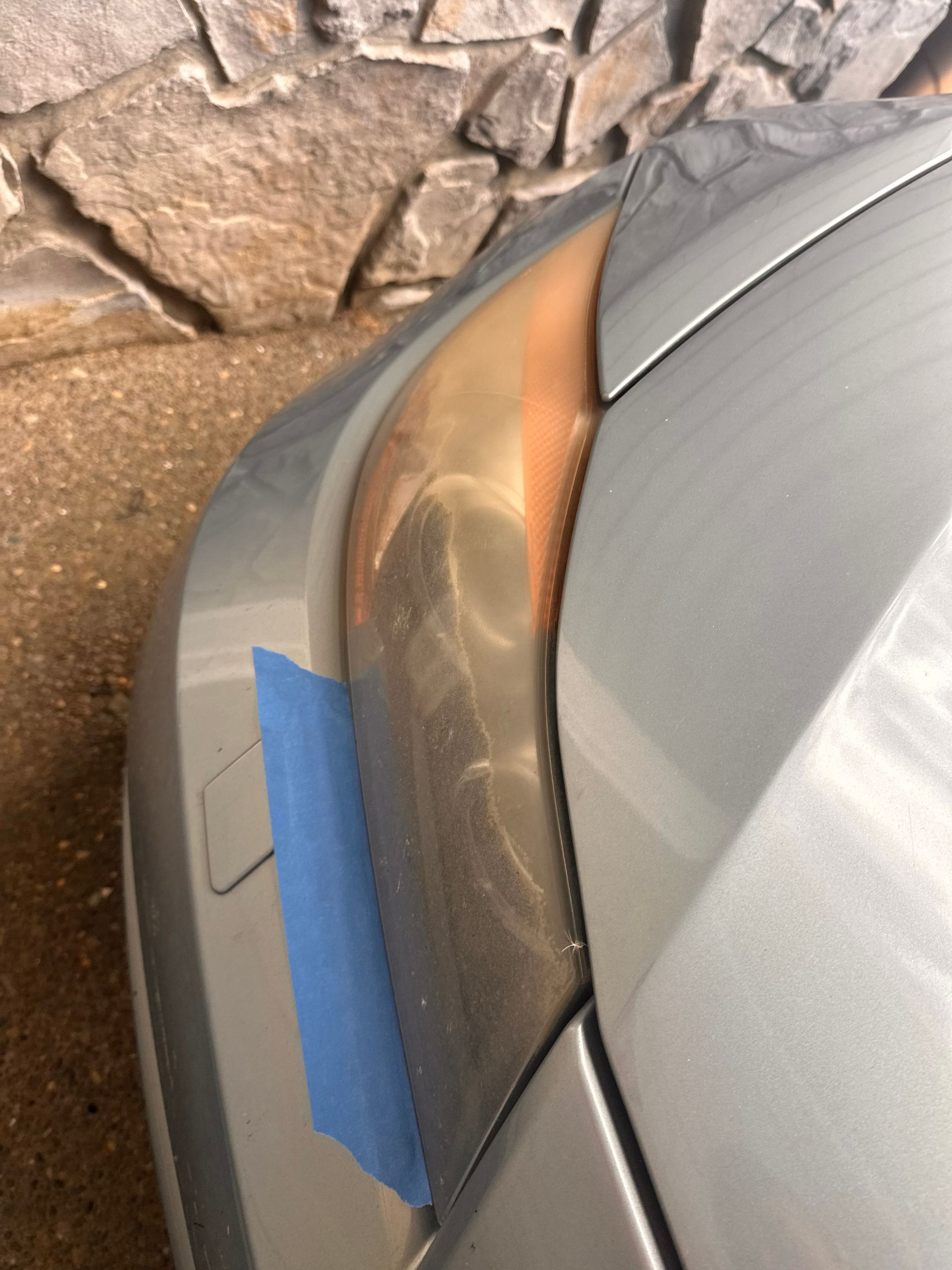 Close-up of a gray car bumper with blue tape covering a scratch near a stone wall