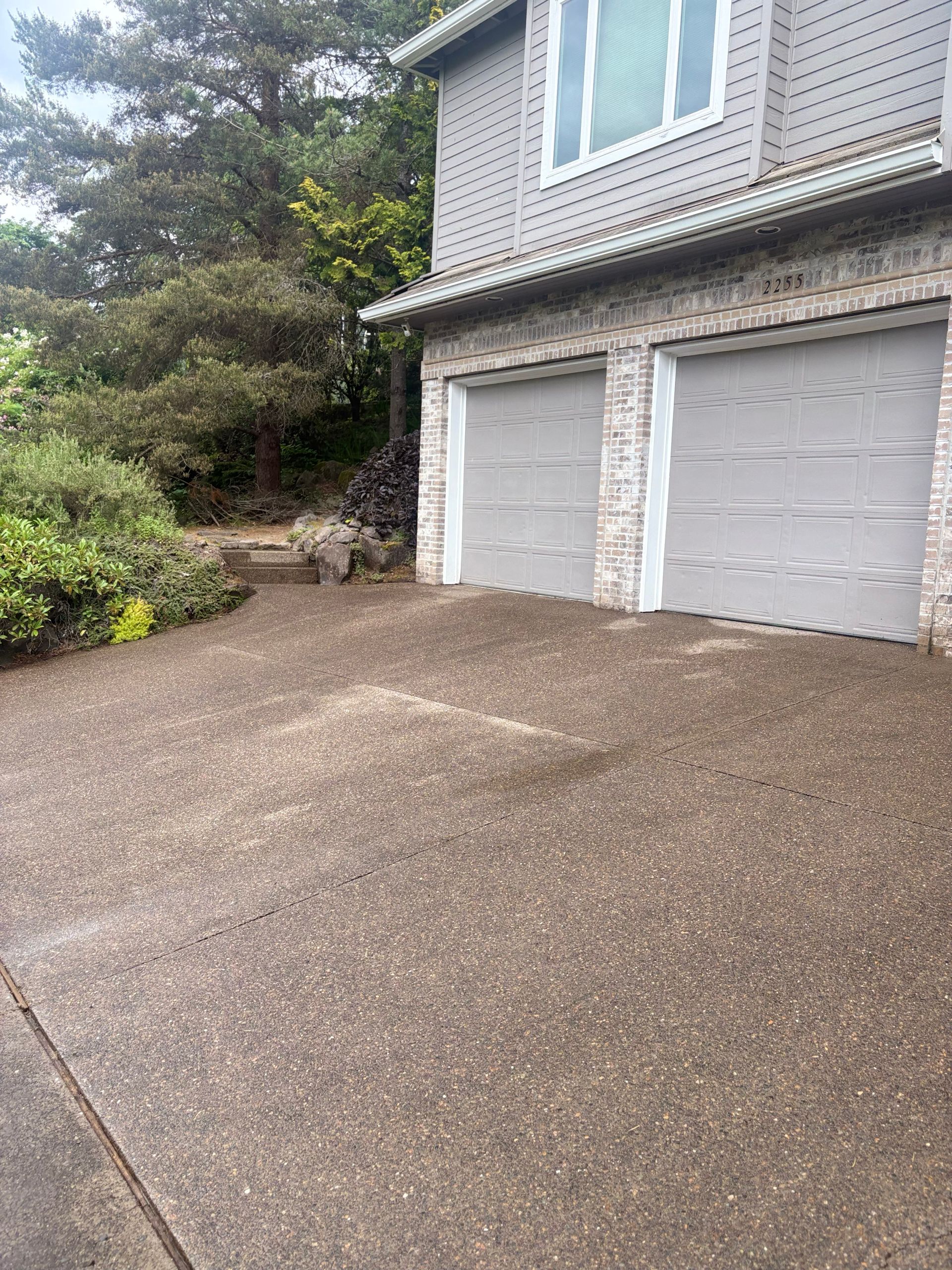 Driveway leading to a two-car garage beside a gray house, with trees and shrubs on the left.