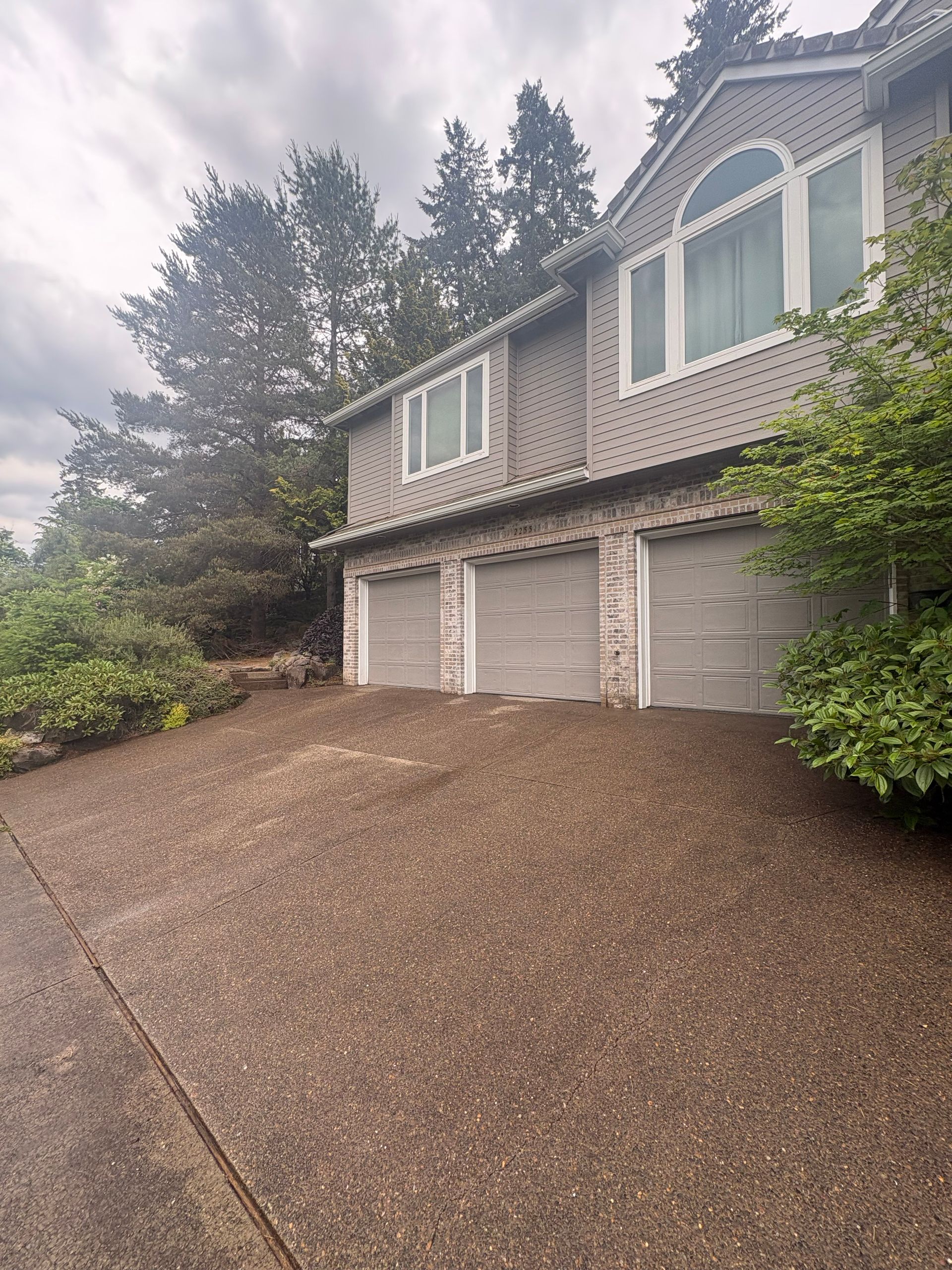 Suburban house with two-car garage, steep gravel driveway, and trees on a cloudy day