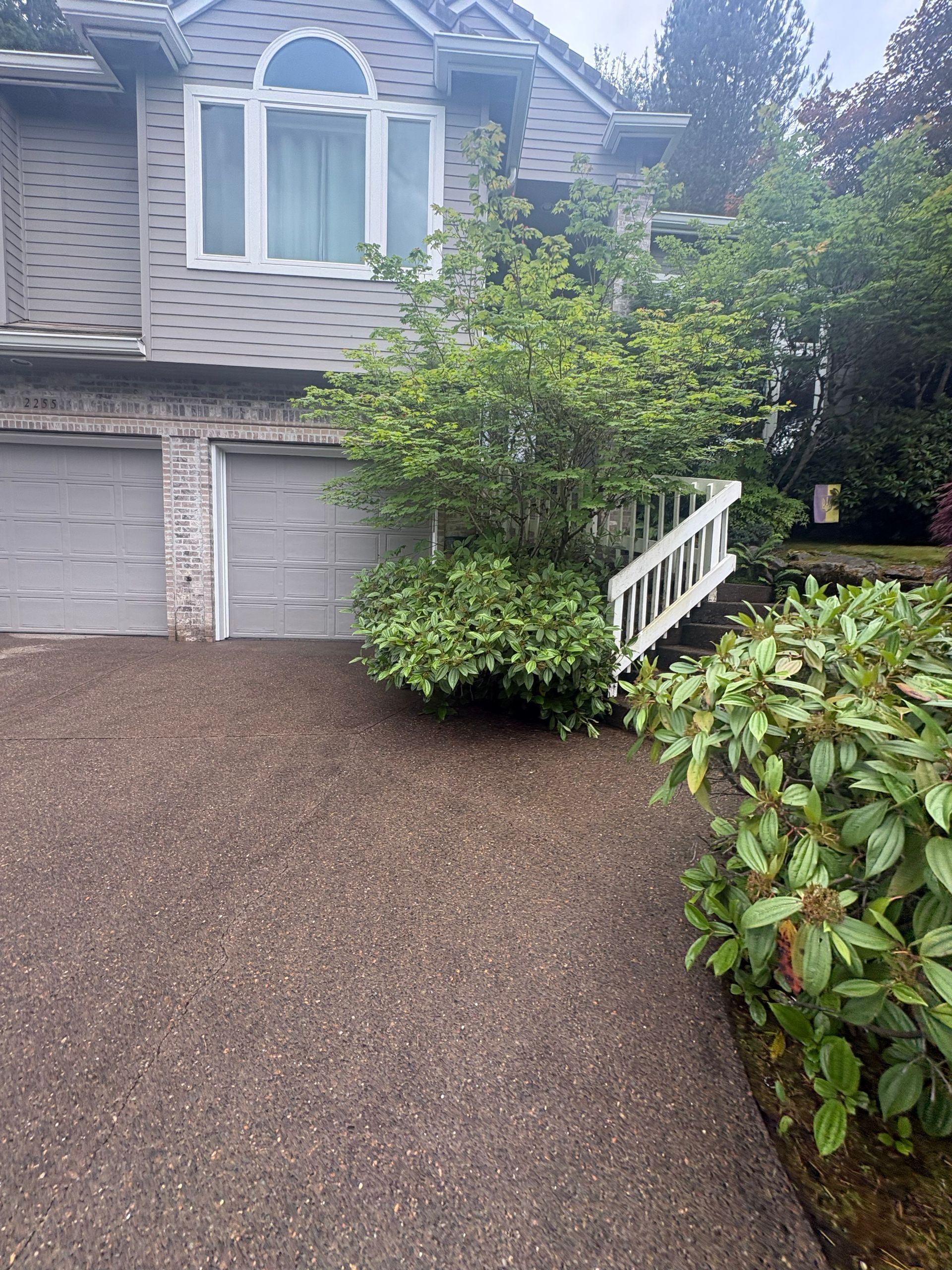 Brick house with garage, gravel driveway, and white railing beside bushes and trees