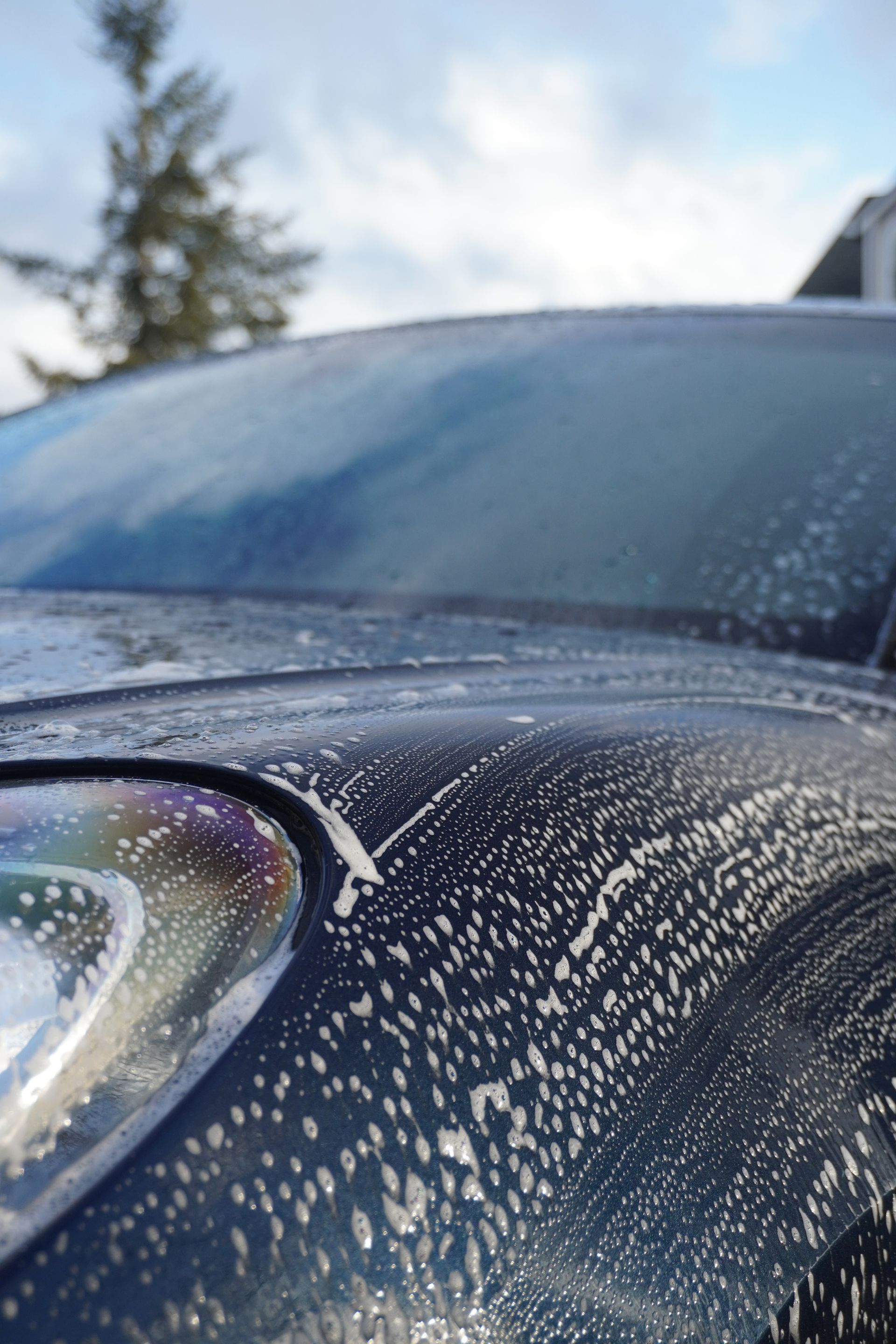 Close-up of a dark car hood and windshield covered in water droplets under a cloudy sky