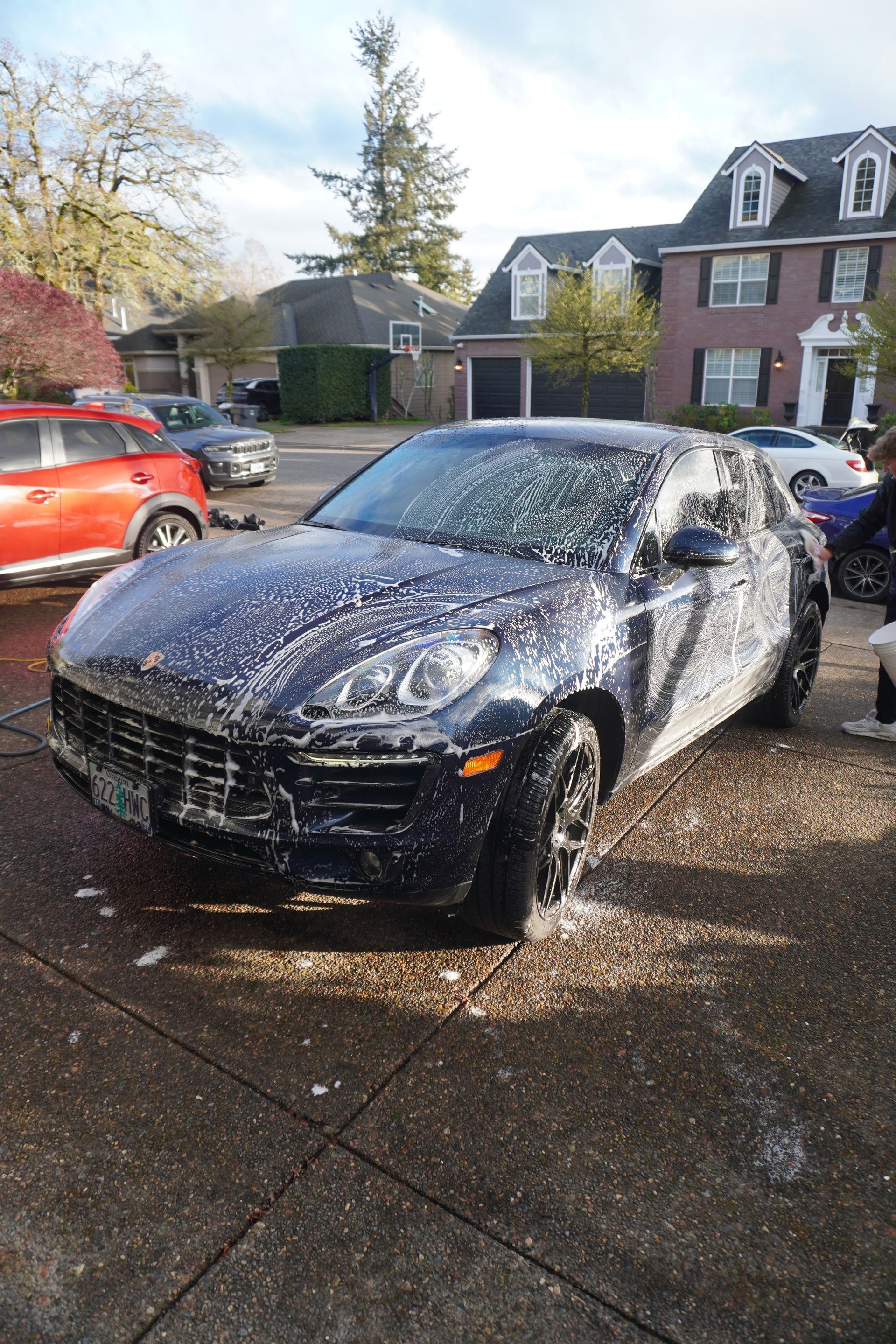 Black car covered in white foam being washed in a driveway, with houses and parked cars nearby