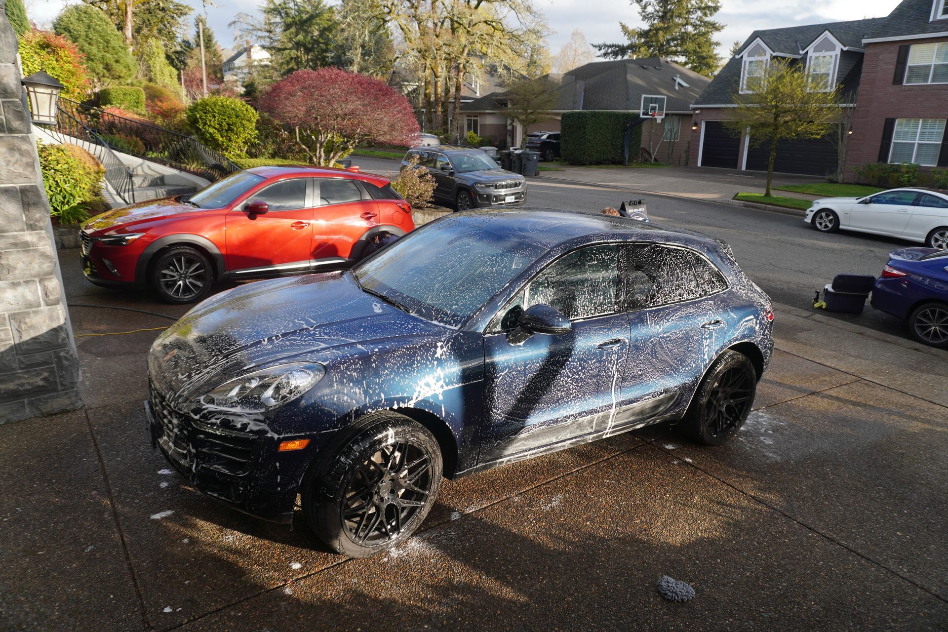 Blue hatchback covered in soapy water being washed on a residential street, with a red car nearby