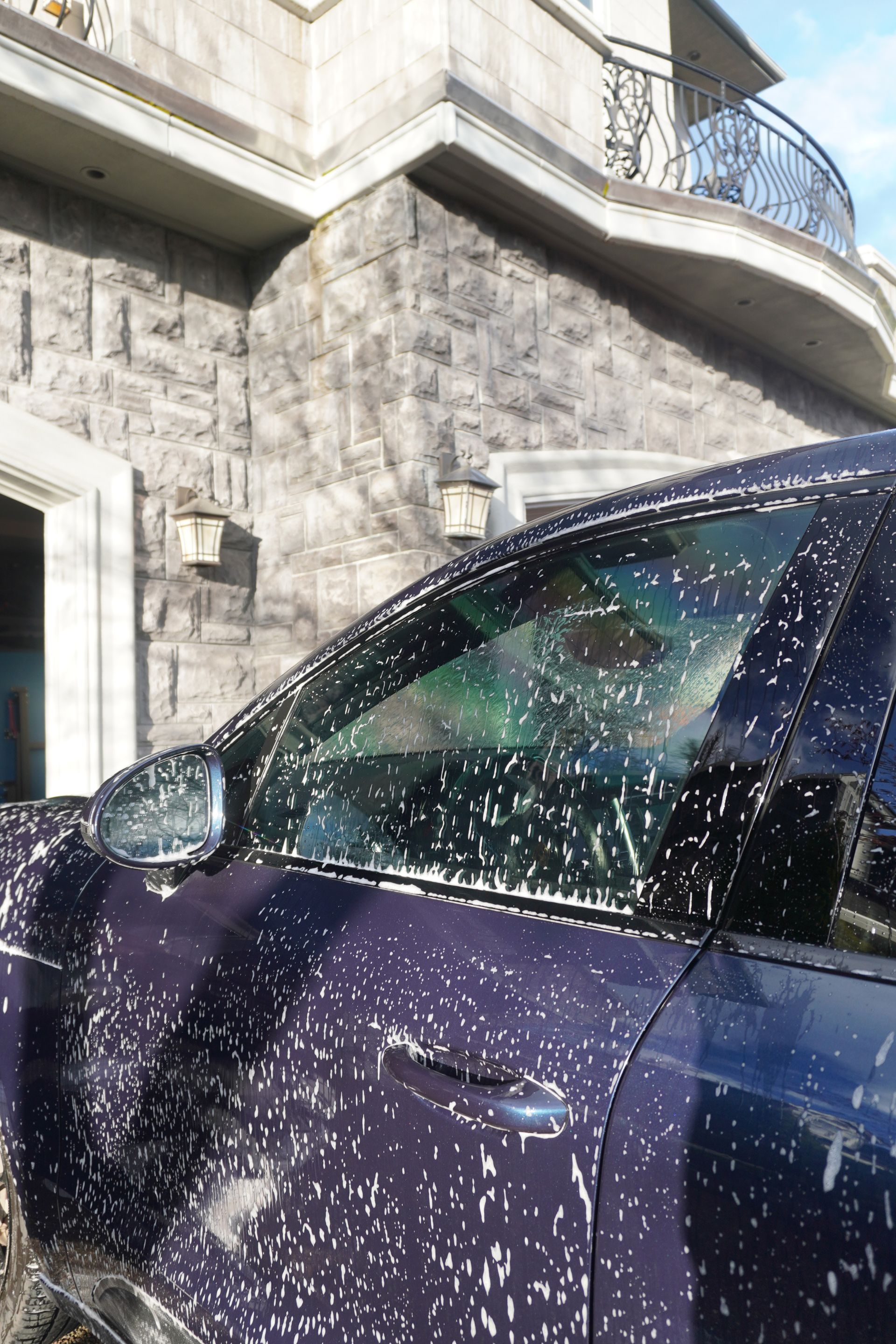 Purple car covered in white soap suds outside a stone building, likely during a car wash.