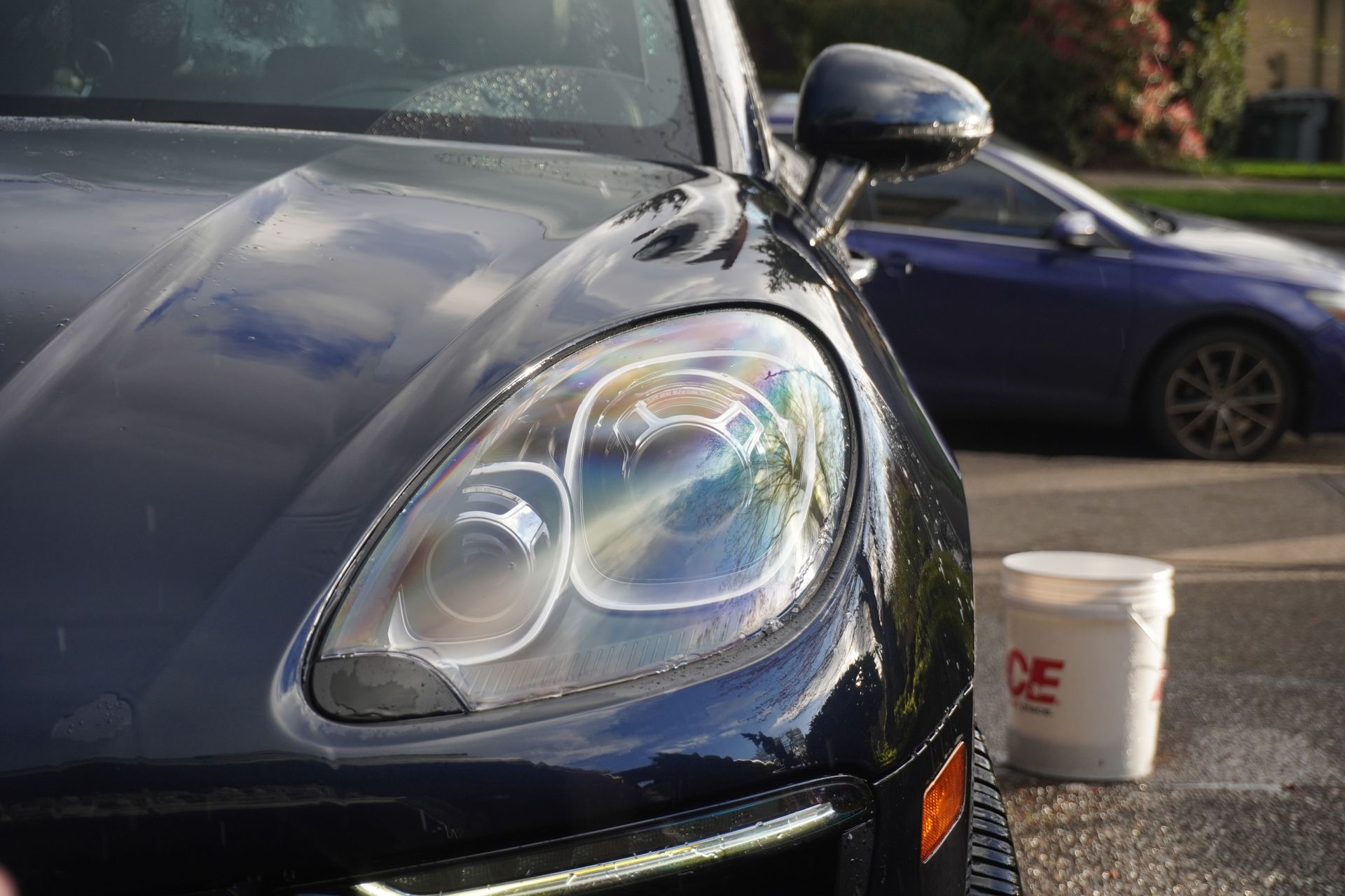 Close-up of a black car's headlight and hood in a parking lot, with a white bucket on the ground nearby