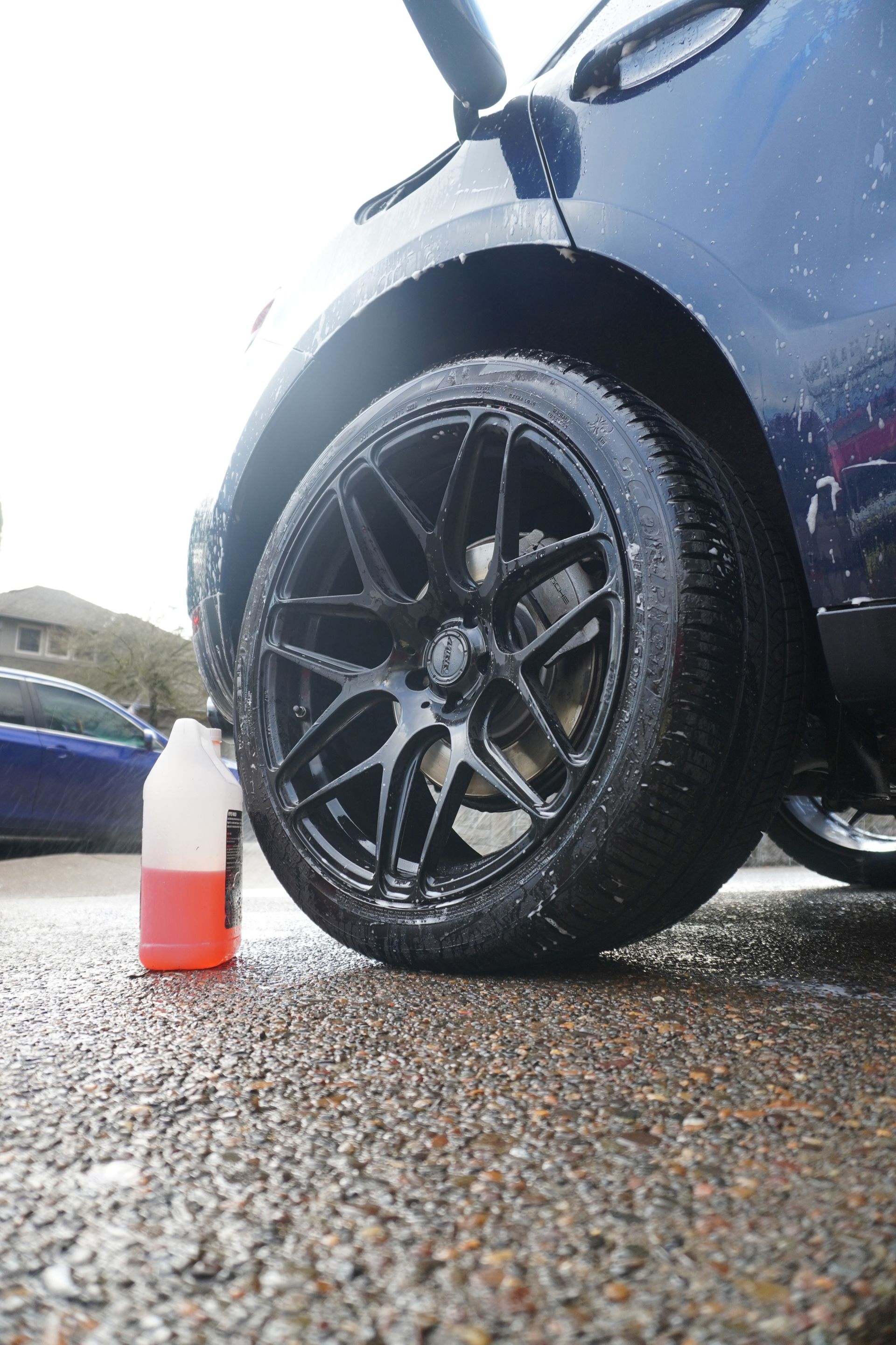Close-up of a car wheel being cleaned with a bottle of red and white solution on wet pavement