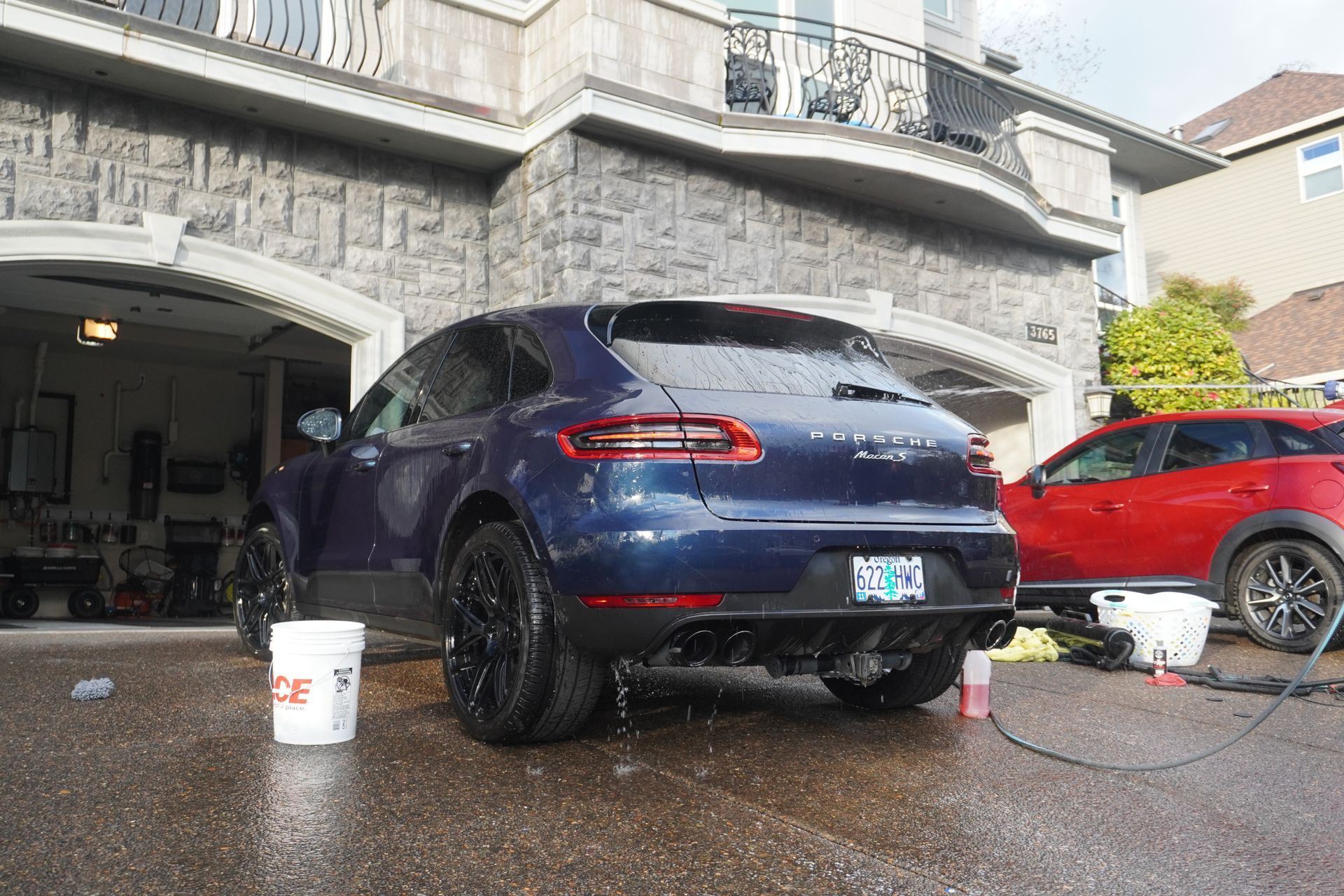 Blue SUV being washed in a driveway, with soap, hoses, and a red car nearby.