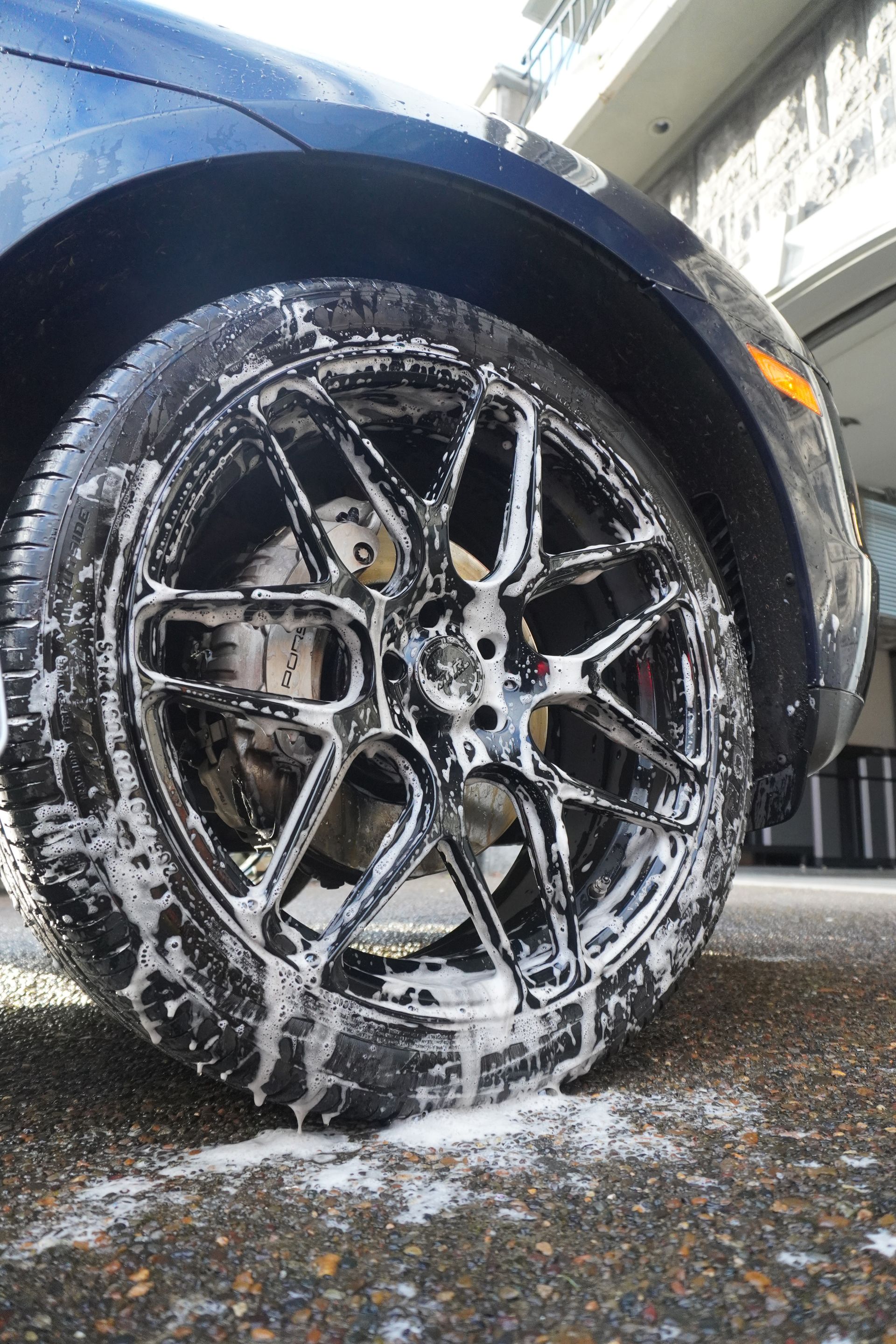 Black car wheel coated in white foam, parked on wet pavement under an overpass.