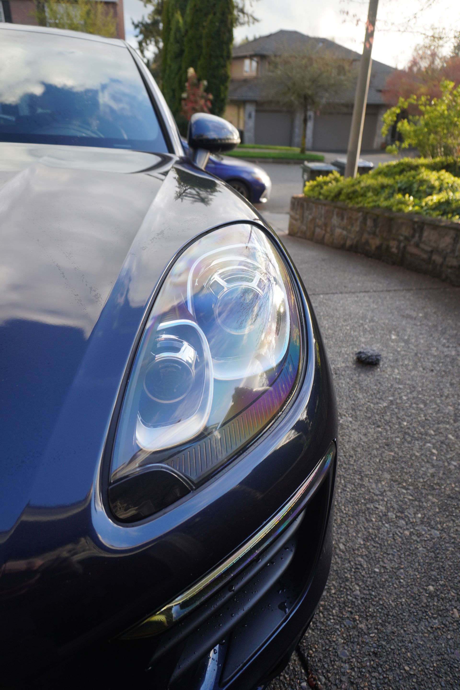 Close-up of a blue car’s front headlight and hood parked on a driveway near a stone wall.