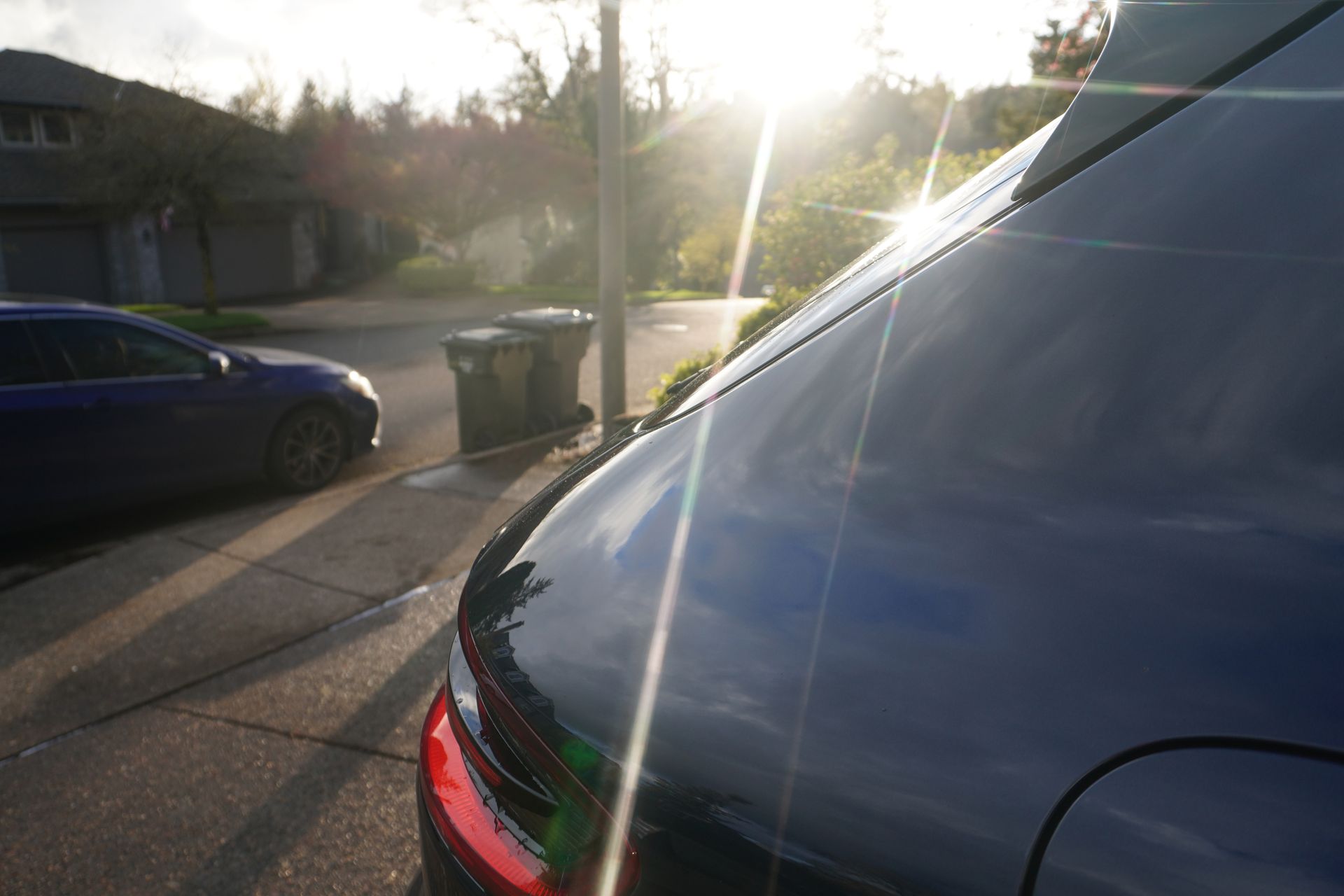 Rear view of a black car on a sunlit residential street with a blue car and trash bins nearby