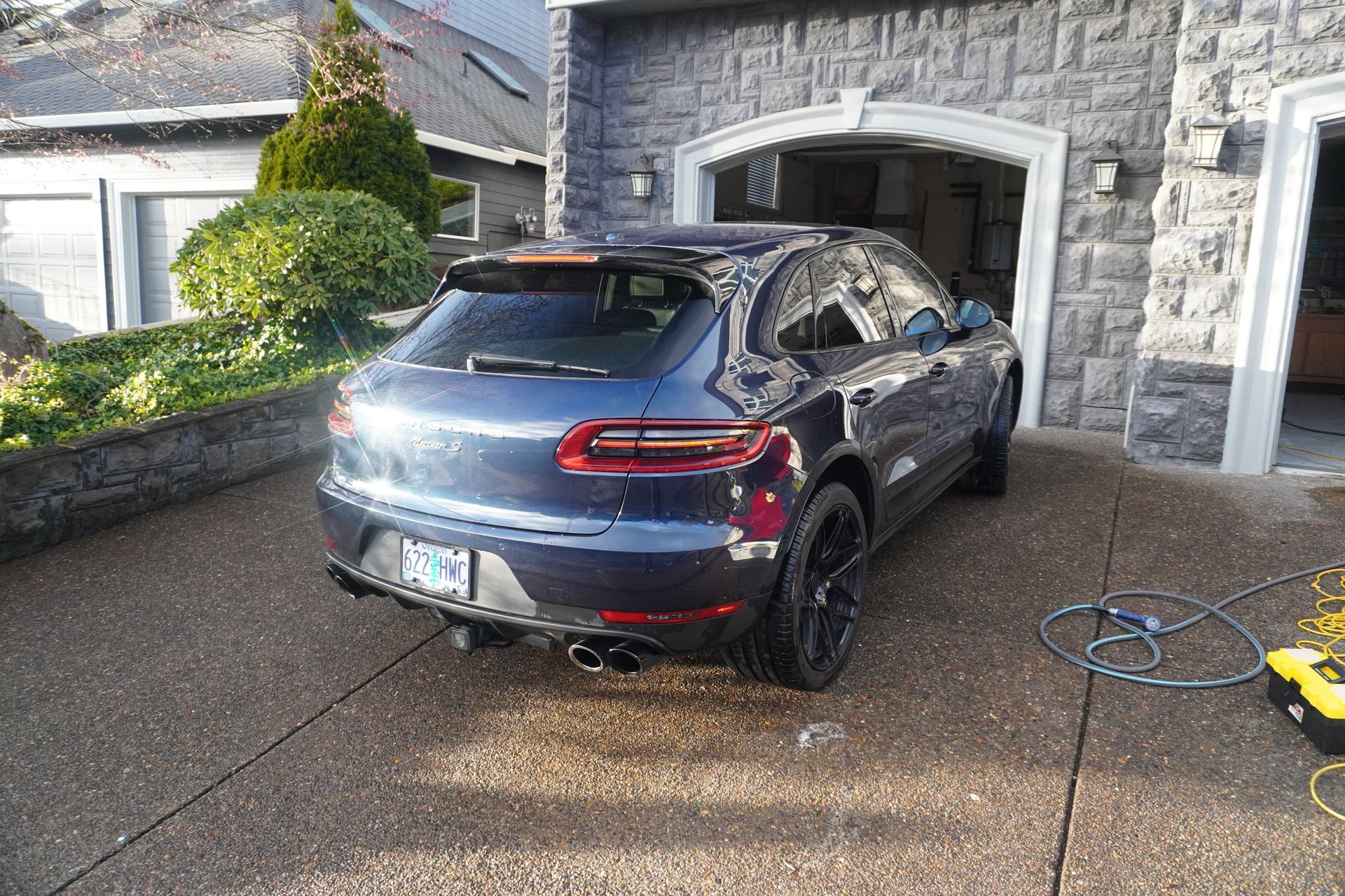 Blue sports SUV being washed in a driveway beside a stone garage, with a hose on the ground.