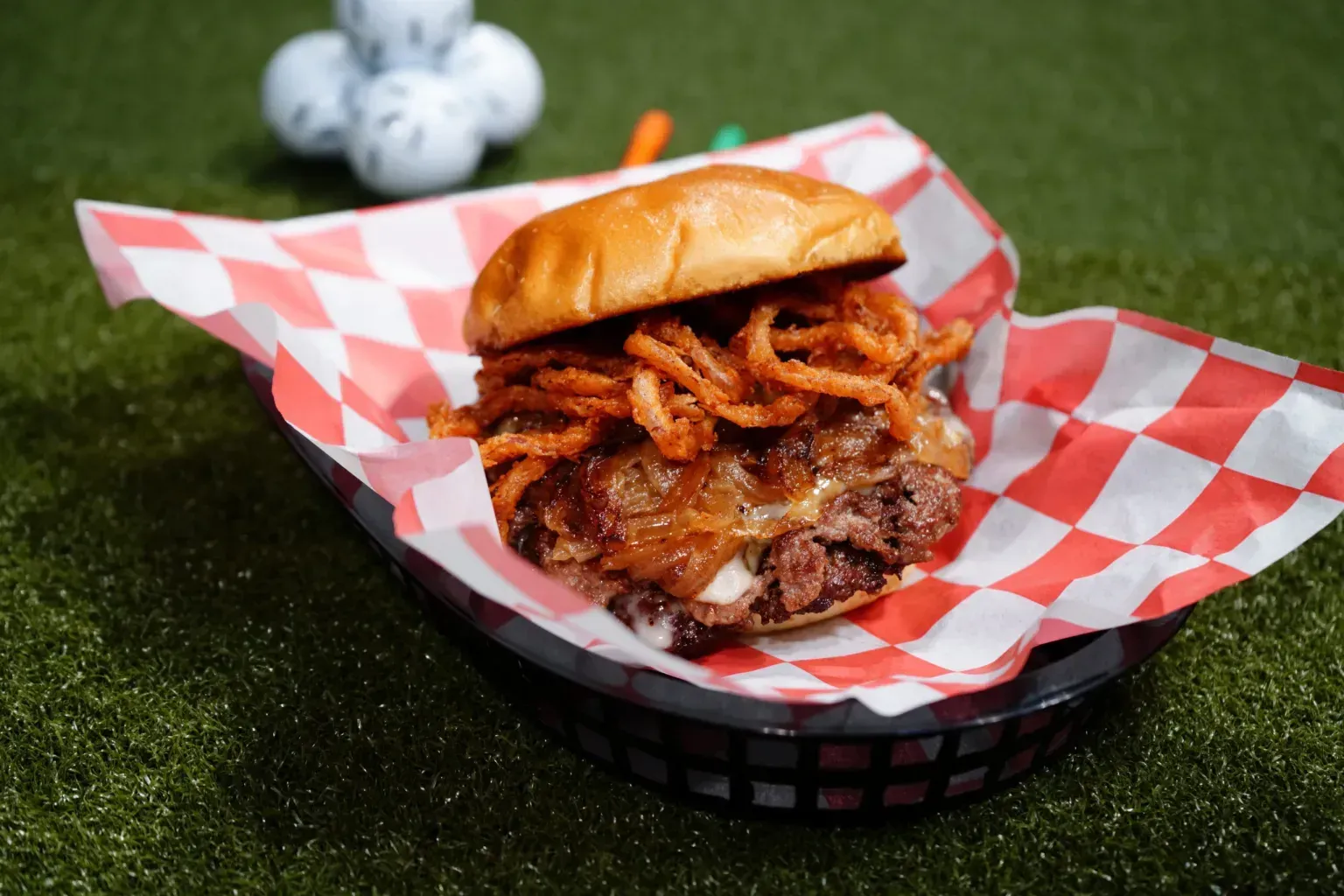 Burger topped with crispy onions in a red-and-white check basket on grass