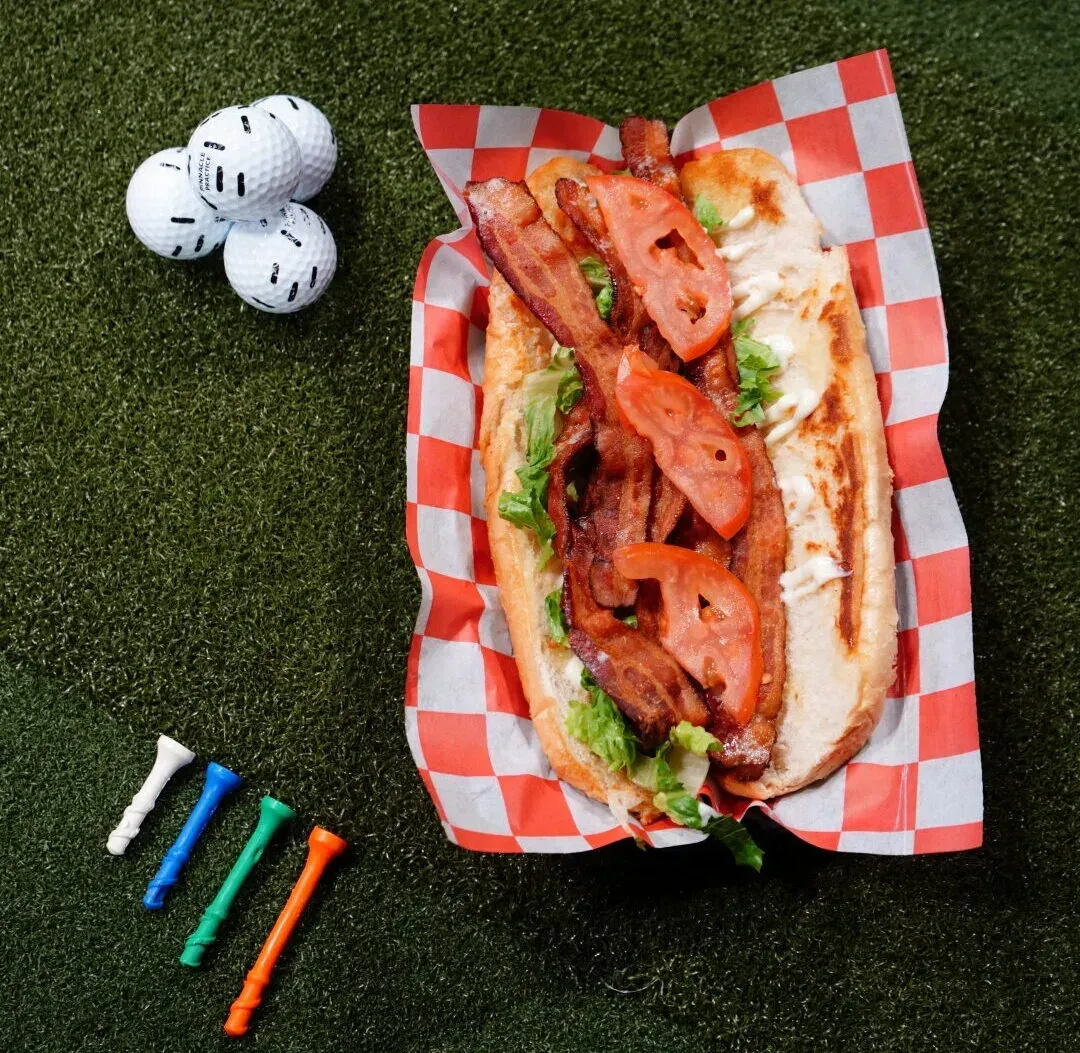 Hot dog with bacon on red-and-white paper, beside three golf balls and four colored tees on grass