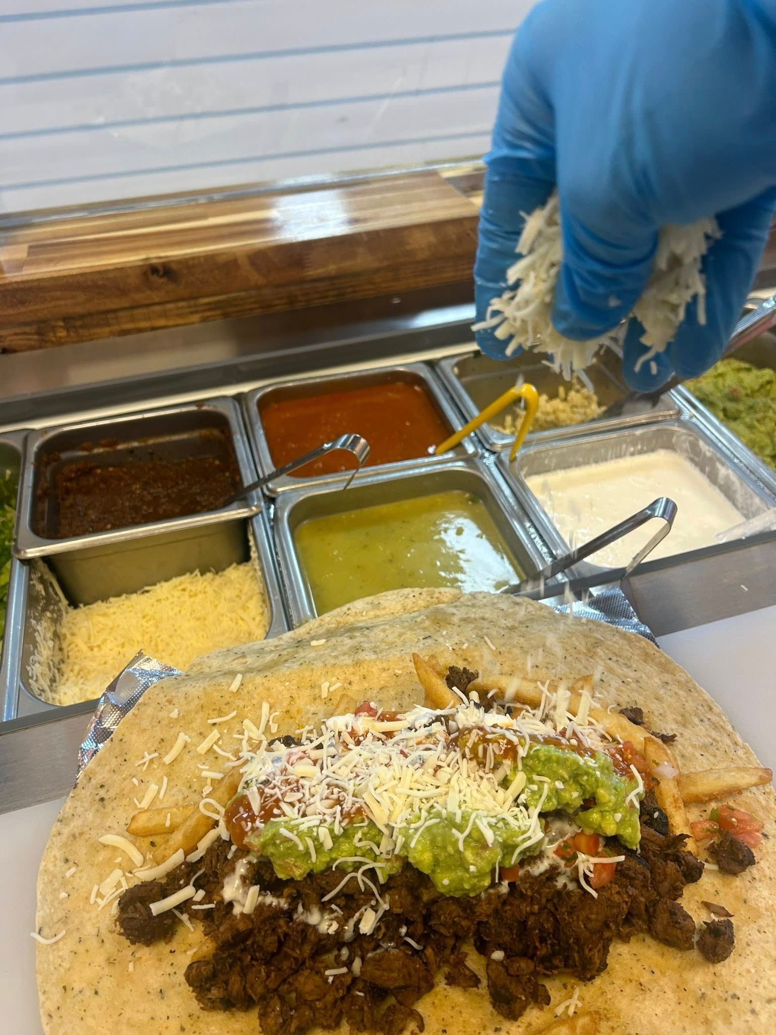 Person in a blue glove sprinkles cheese onto a burrito with fillings like guacamole and meat, near various toppings.