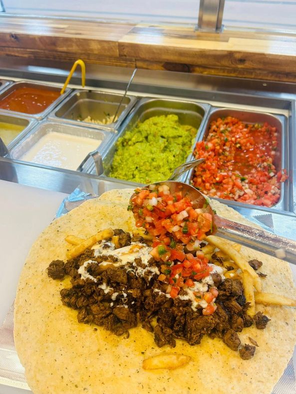 Burrito being assembled with toppings: salsa, guacamole, and sauces at a food station.