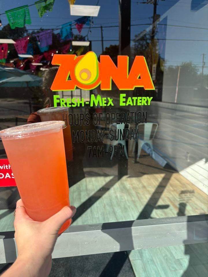 Hand holding a large, pink drink in front of the Zona Fresh-Mex Eatery storefront. The sign has an avocado logo.