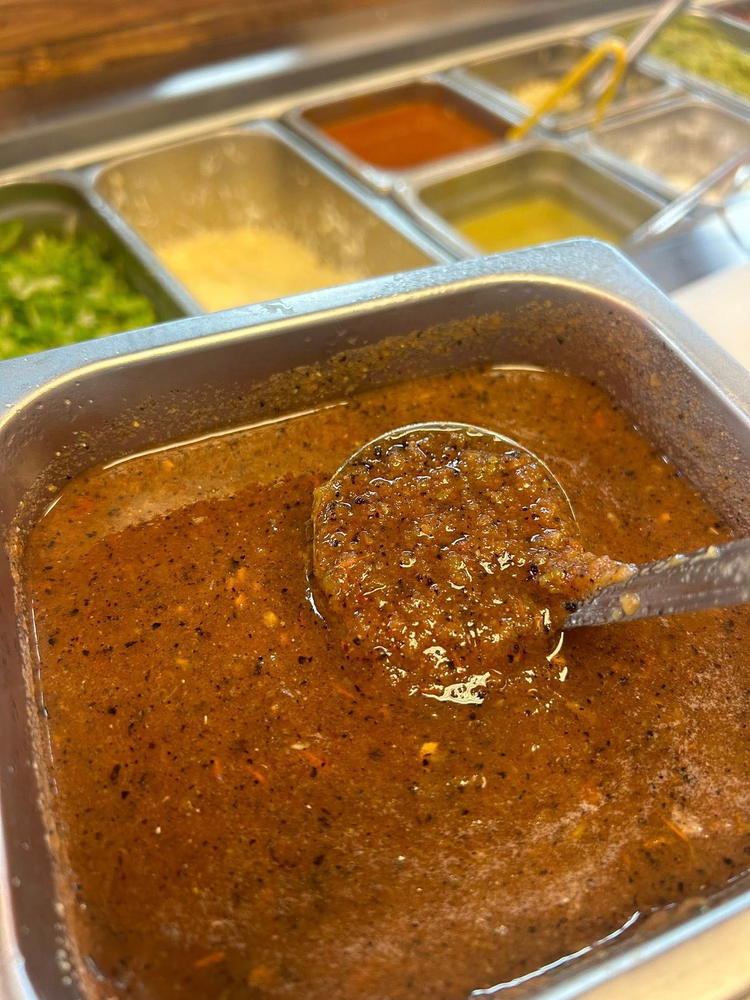 A metal container of salsa with a ladle inside. Other ingredients are visible in the background.