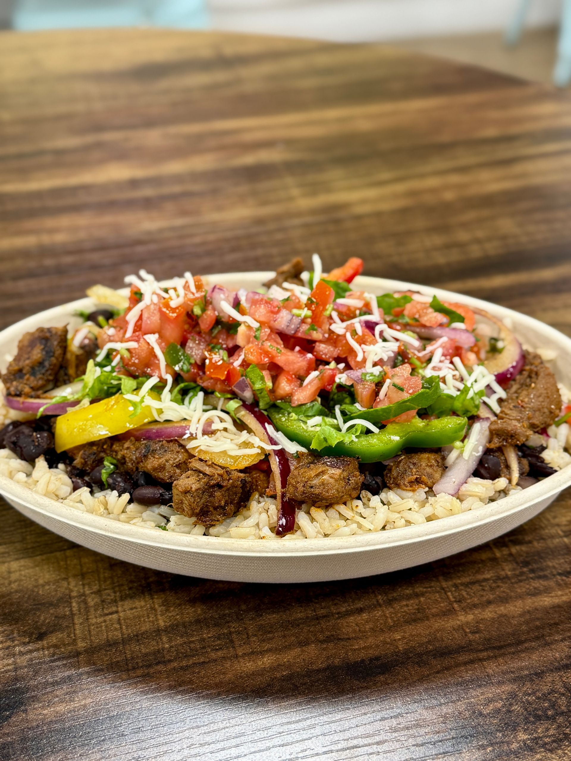 Burrito bowl with brown rice, black beans, meat, vegetables, and toppings in a beige bowl on wood.