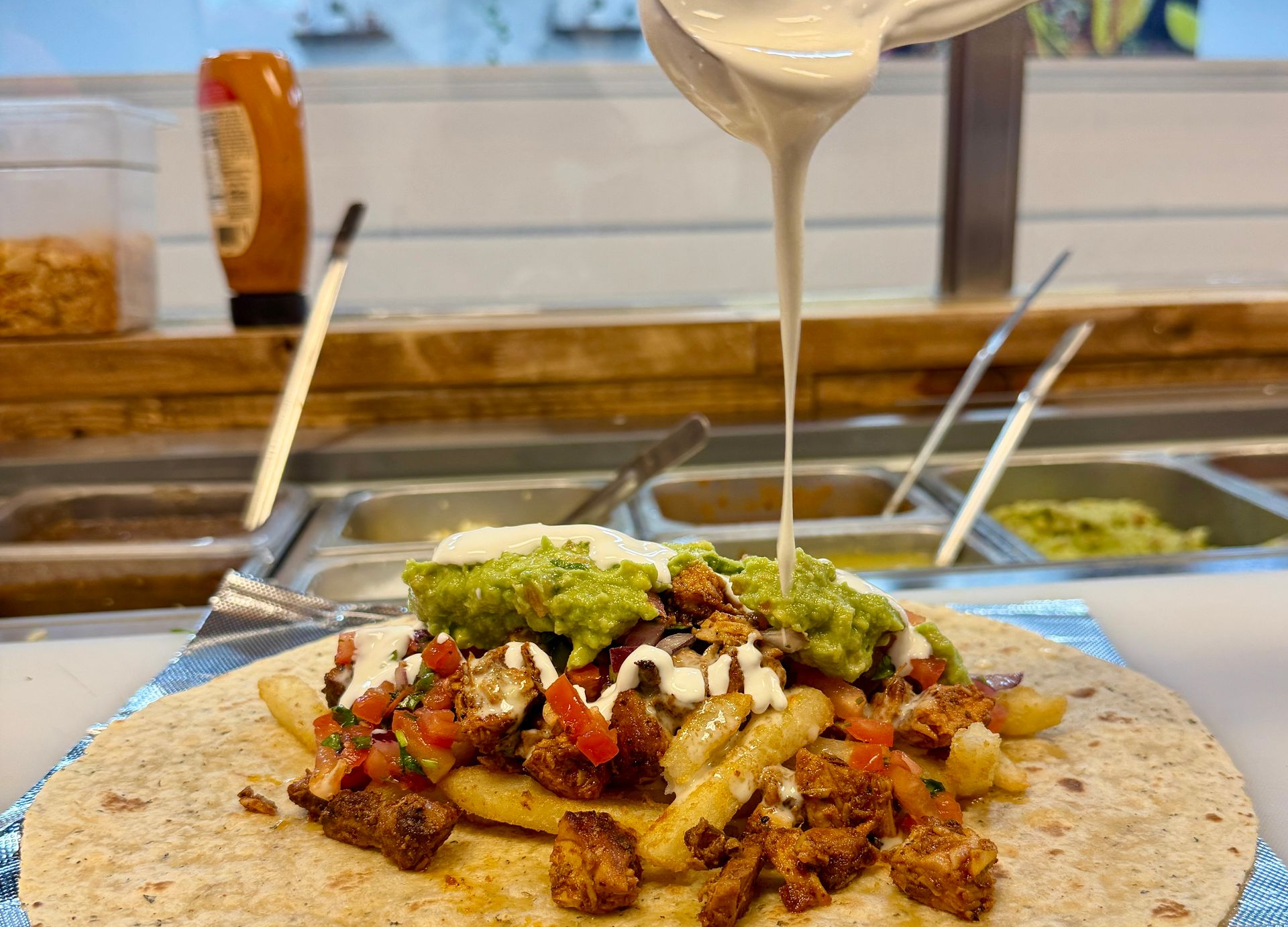 Burrito being drizzled with sauce; contains fries, guacamole, and other fillings on a tortilla.