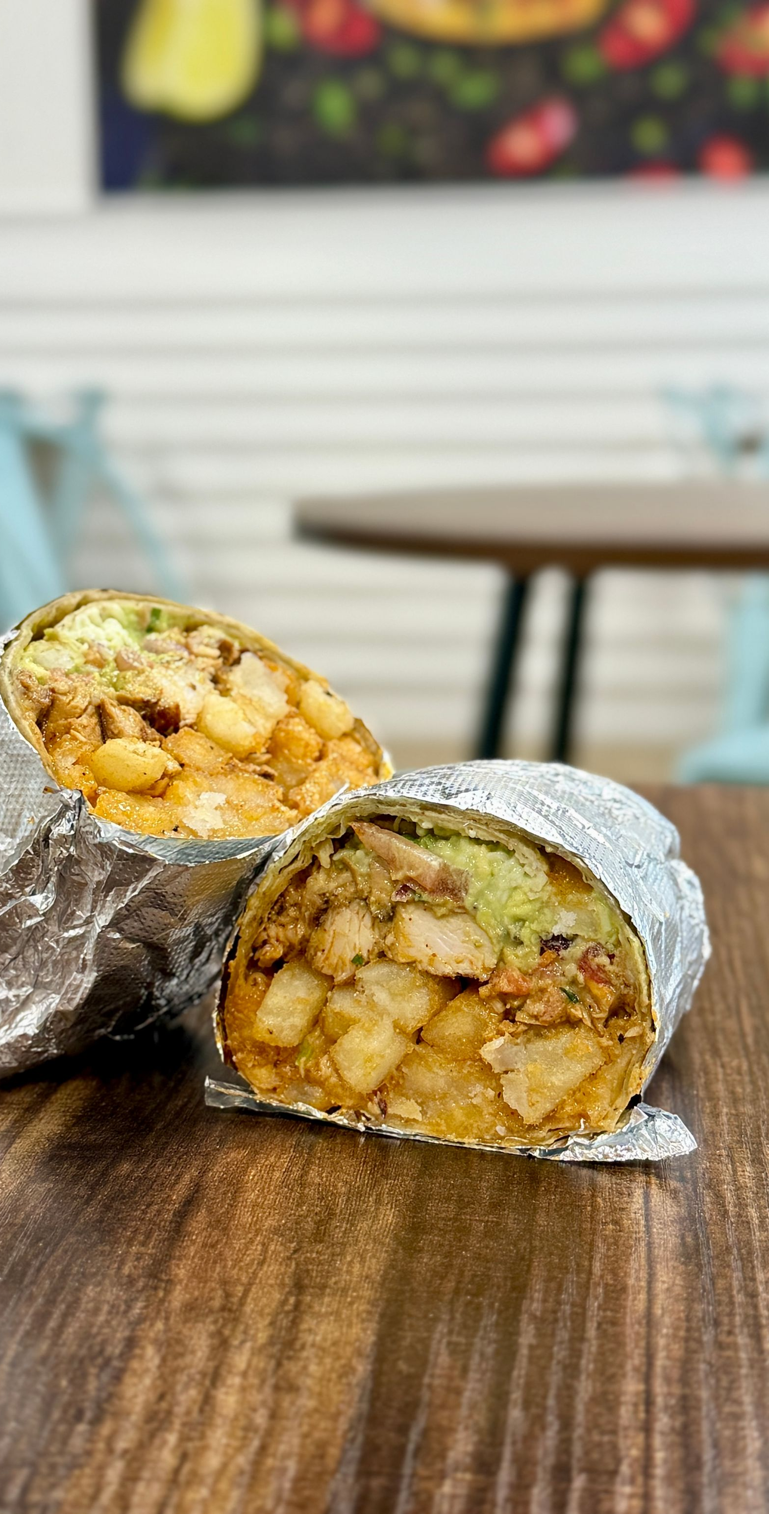Burrito wrapped in foil cut in half, revealing fried potatoes, meat, and avocado, on a wood table.