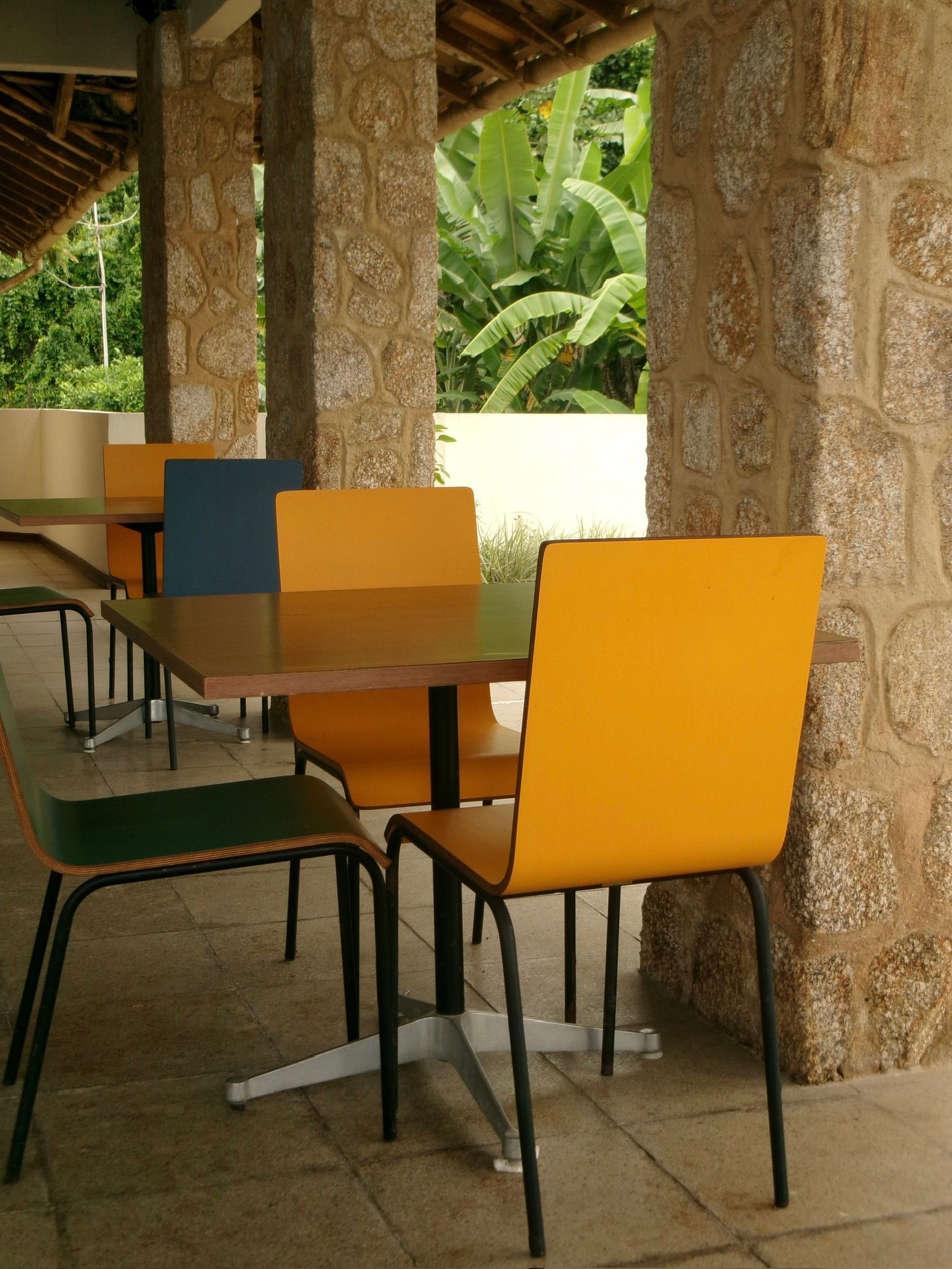 Mesas vazias de um café ao ar livre com cadeiras laranja e azul sob um pátio coberto.