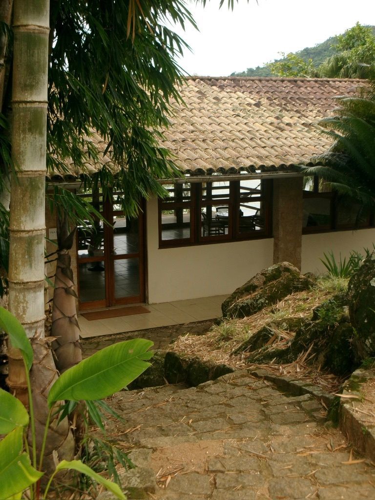 Caminho de pedra que leva a uma casa rústica com telhado de telhas, emoldurada por plantas e árvores tropicais.
